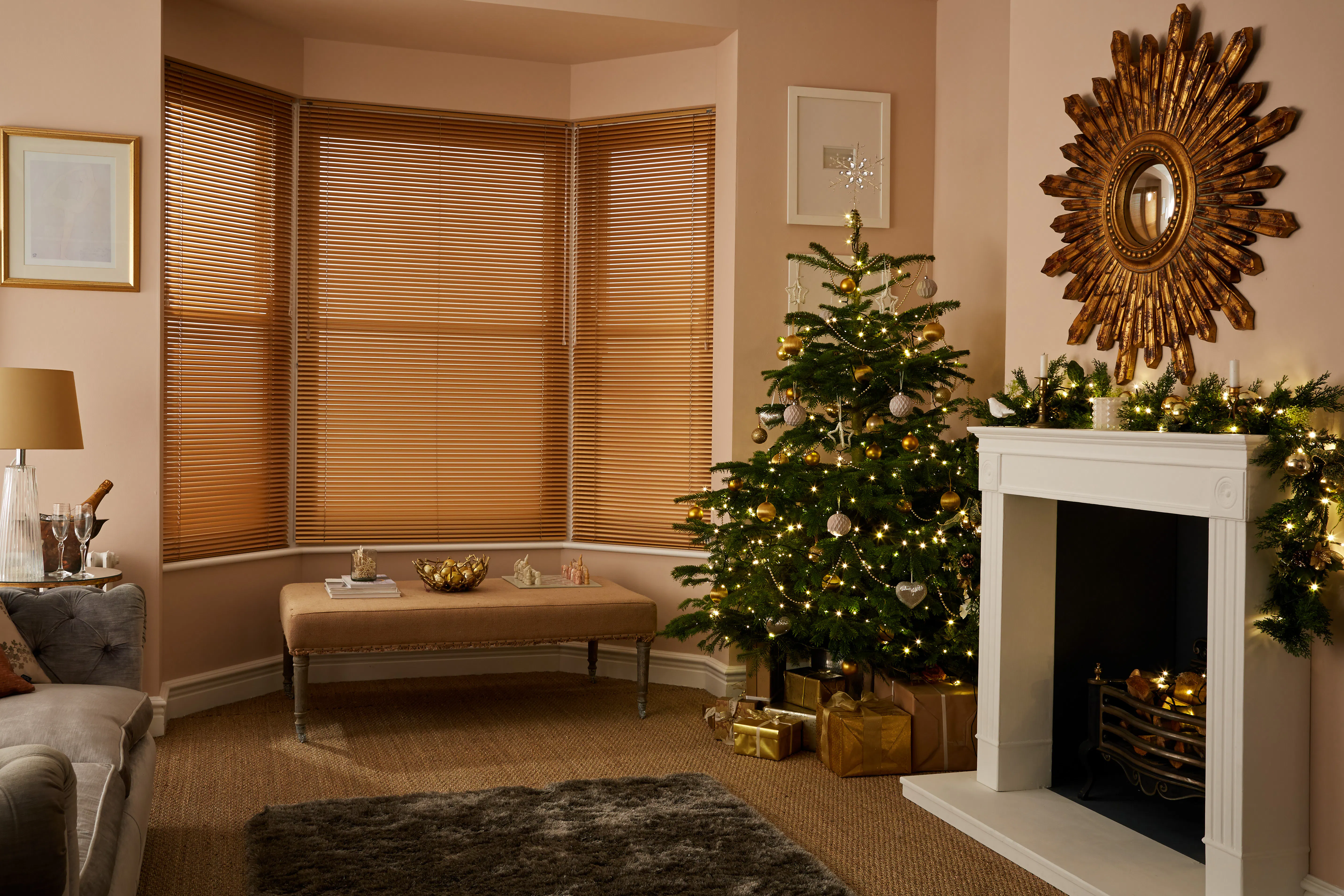 Bay window with warm gold Venetian blinds beside a decorated Christmas tree, creating a cosy living room scene with a fireplace.
