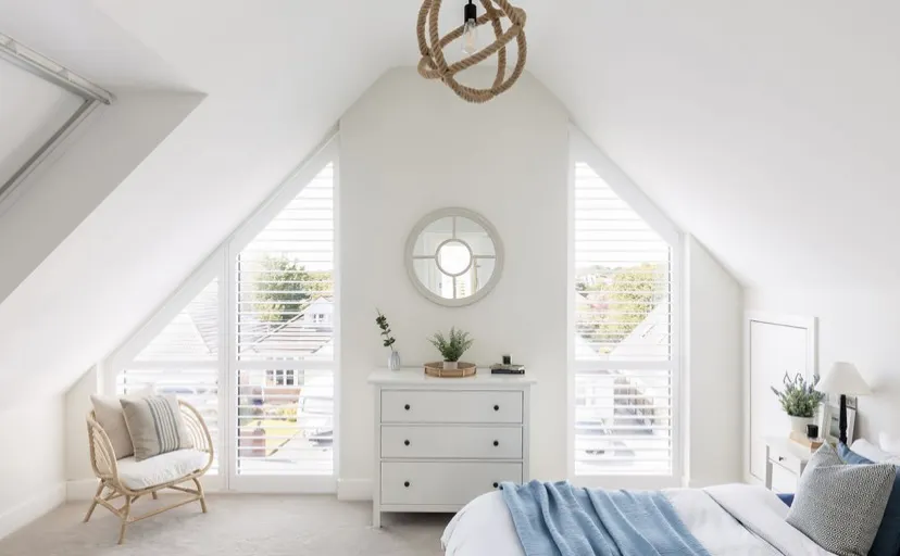 A bright, all-white loft bedroom with white shutters on angled apex windows, a rattan chair and a rope pendant light.