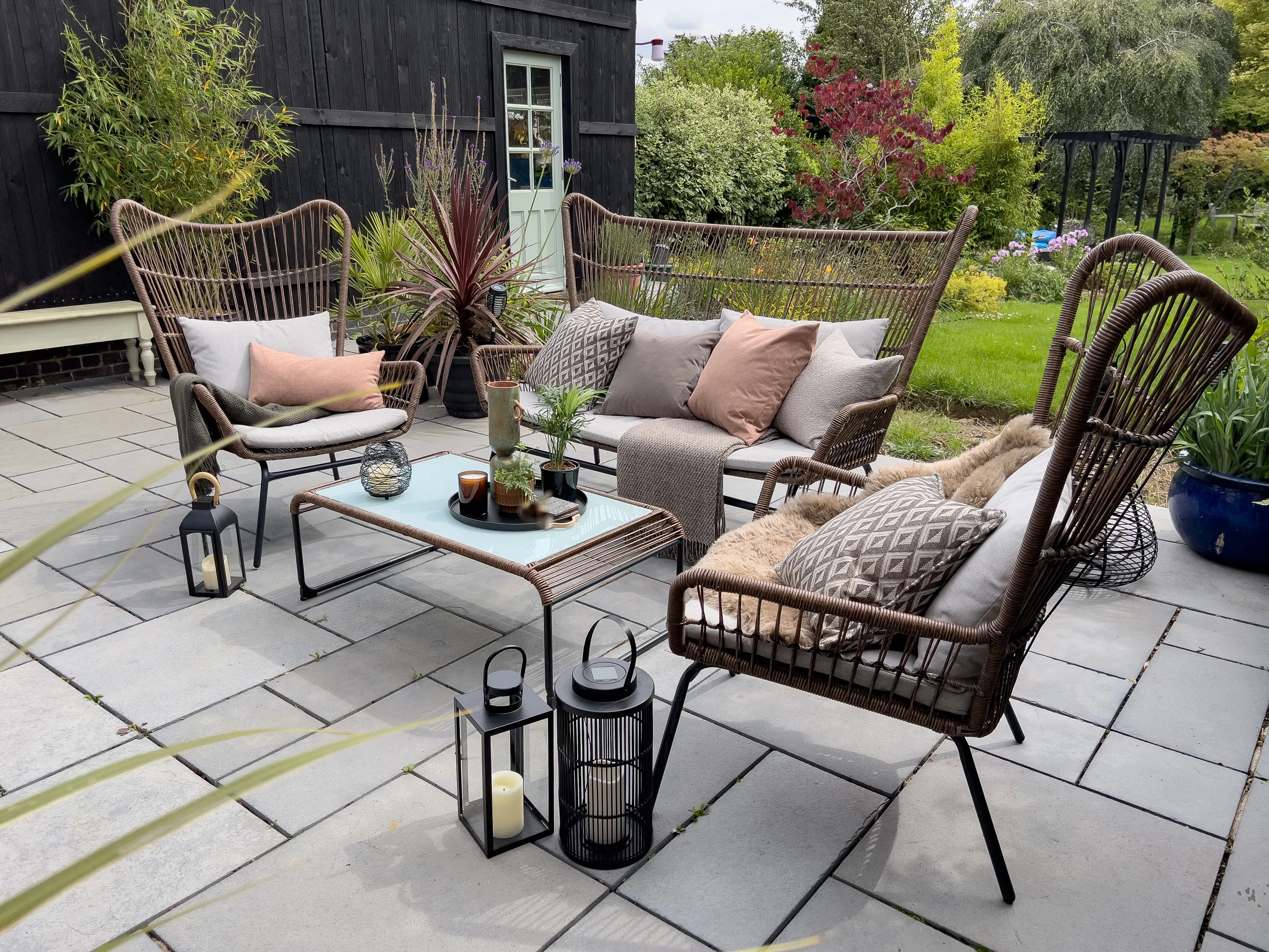 Rattan garden sofa set with blush and grey cushions on a stone patio, styled with black lanterns and surrounded by lush planting.