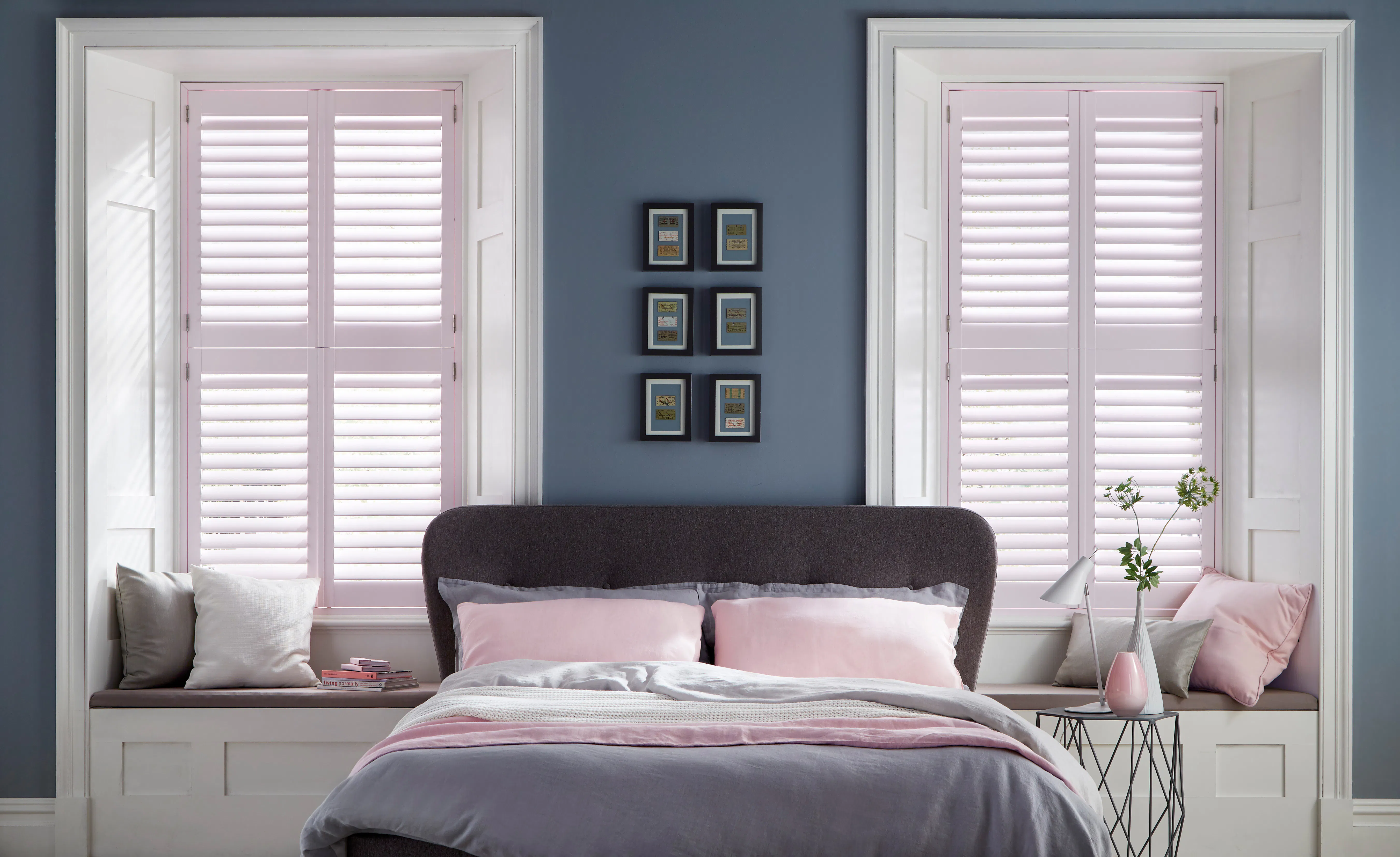 A bedroom with two window seats either side of the bed, each dressed with pale pink plantation shutters and layered cushions.