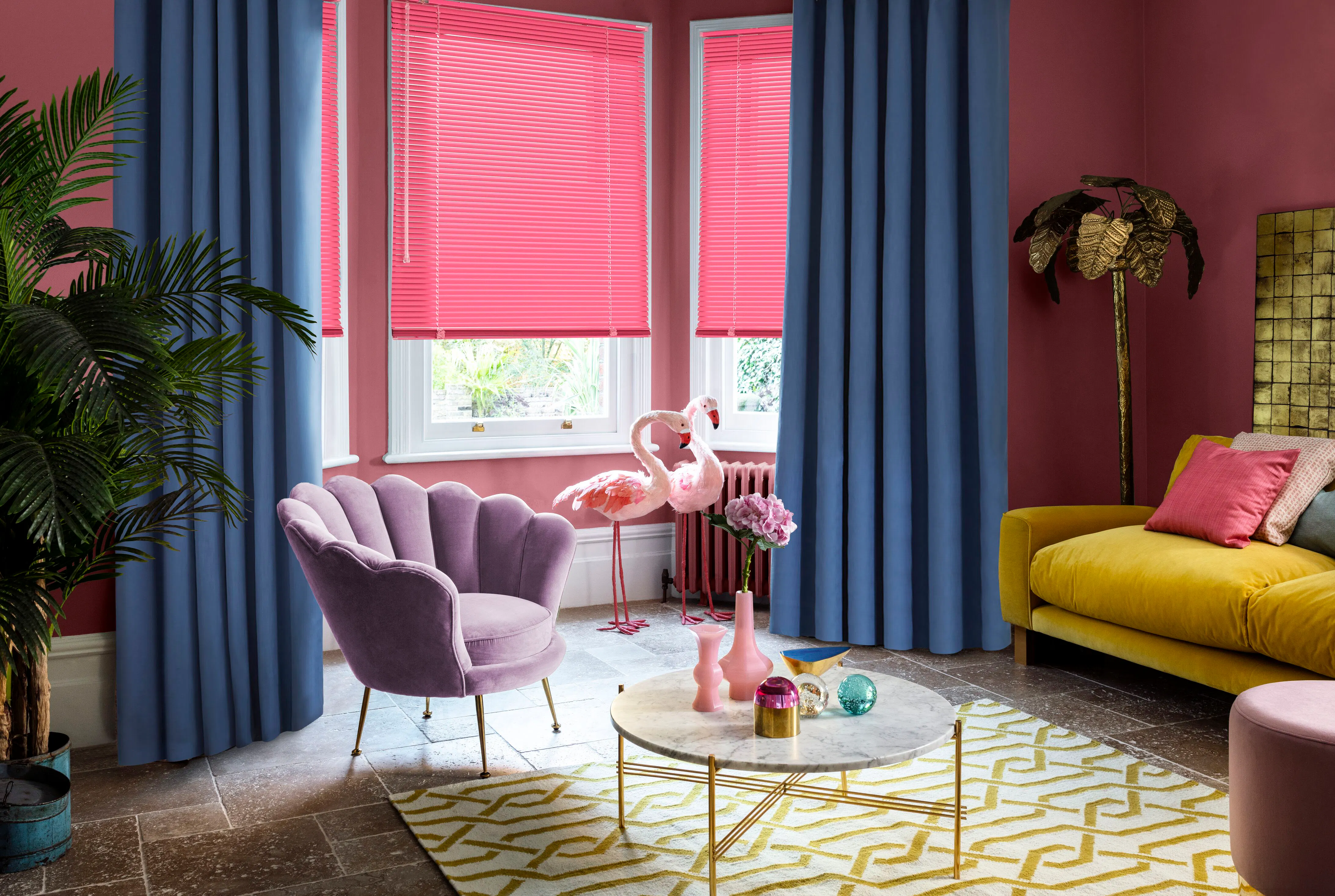 A colourful living room with bright pink walls, blue floor-to-ceiling curtains, a pink Venetian blind and a mustard velvet sofa, styled with pink flamingo ornaments.