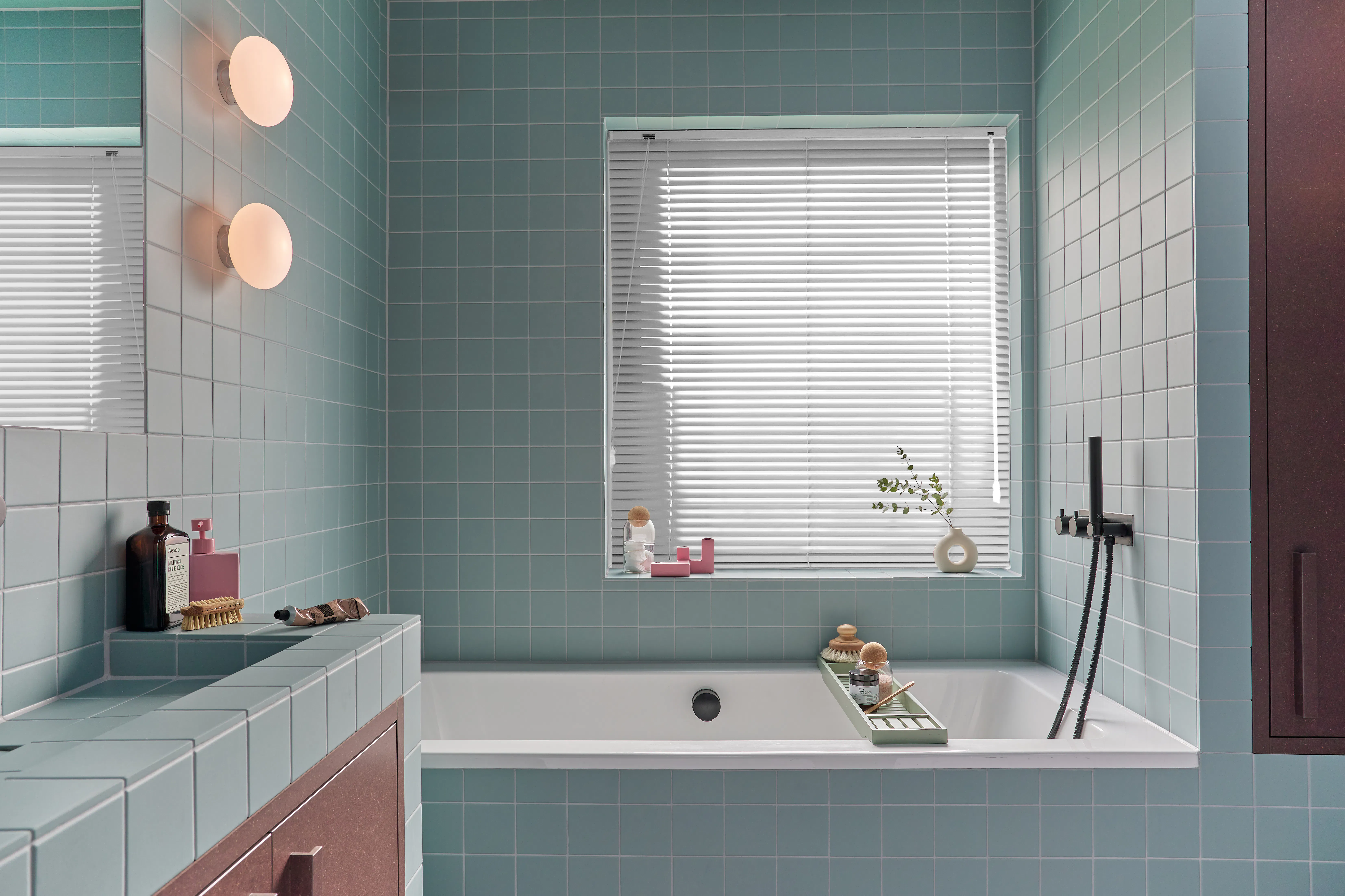 White aluminium venetian blind covering a square bathroom window above a teal‑tiled bathtub with modern black fittings.
