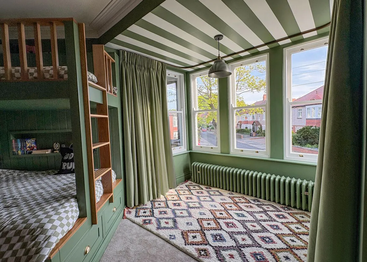 Three bay windows with sage-green curtains bring natural light into a green bedroom, featuring a striped ceiling, radiator and patterned rug.