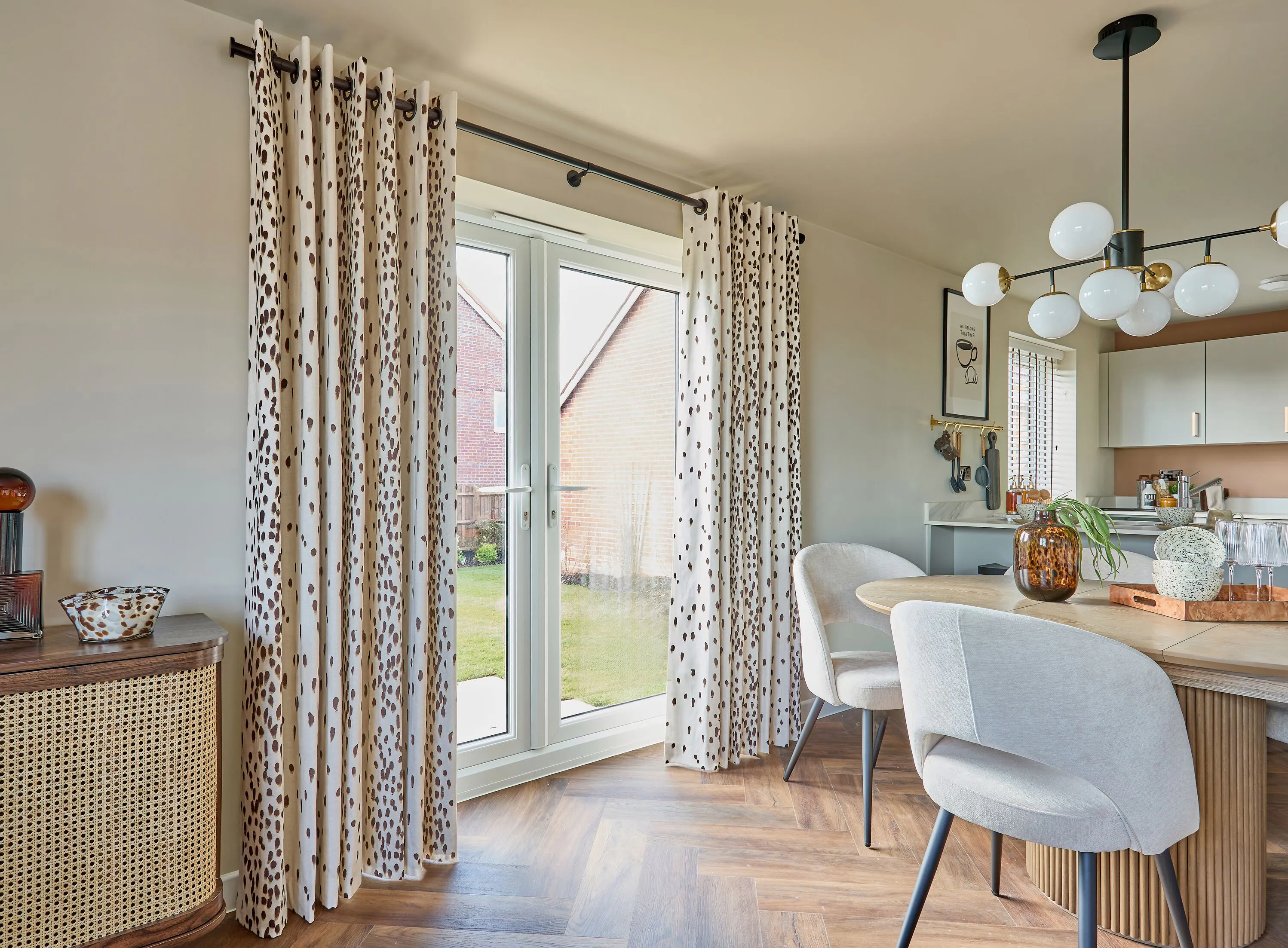 Patterned curtains frame French doors opening to the garden, bringing light into a dining space with a herringbone floor and modern furnishings.