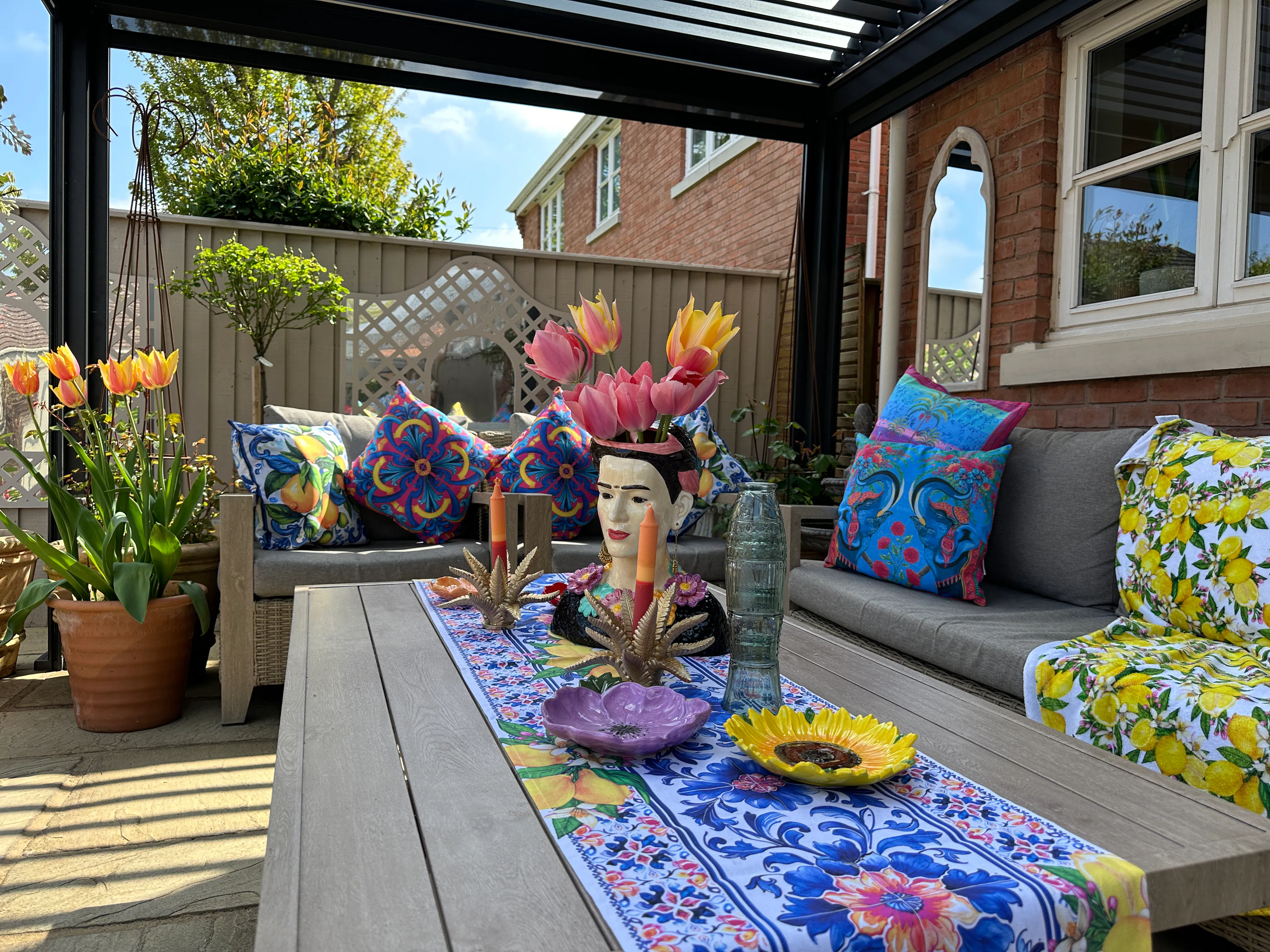 Pergola seating area styled with colourful patterned cushions, a floral table runner and a decorative vase head filled with tulips.