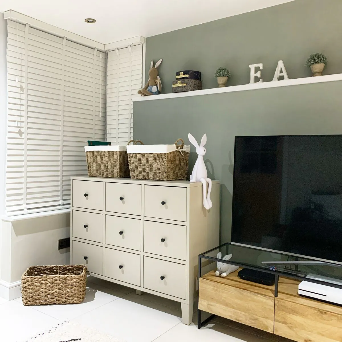 White shuttered corner window beside a storage unit and TV, bringing light into a calm room with sage-green walls and woven baskets.