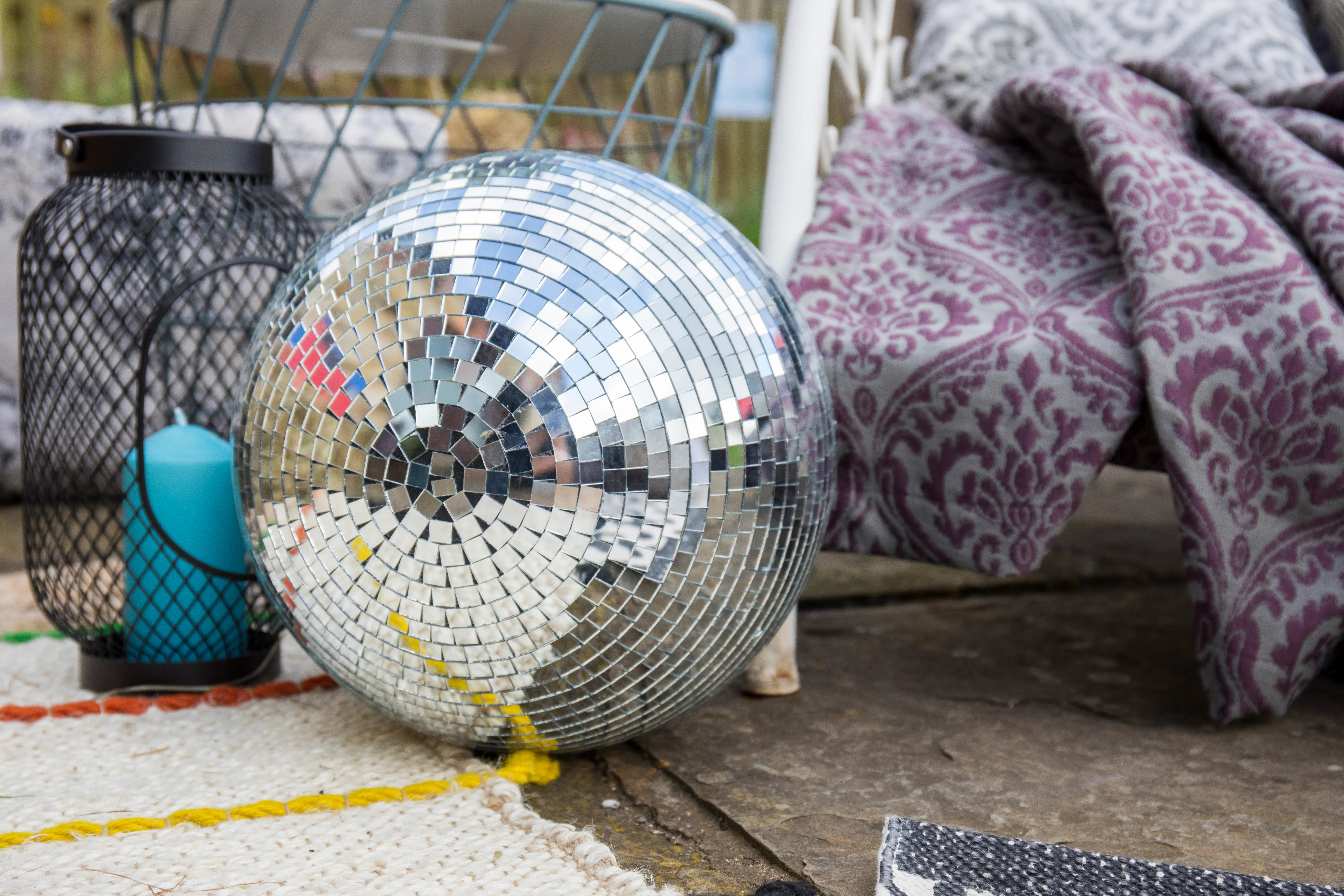 Close-up of a mirrored disco ball beside a lantern and patterned outdoor cushions on a garden patio.