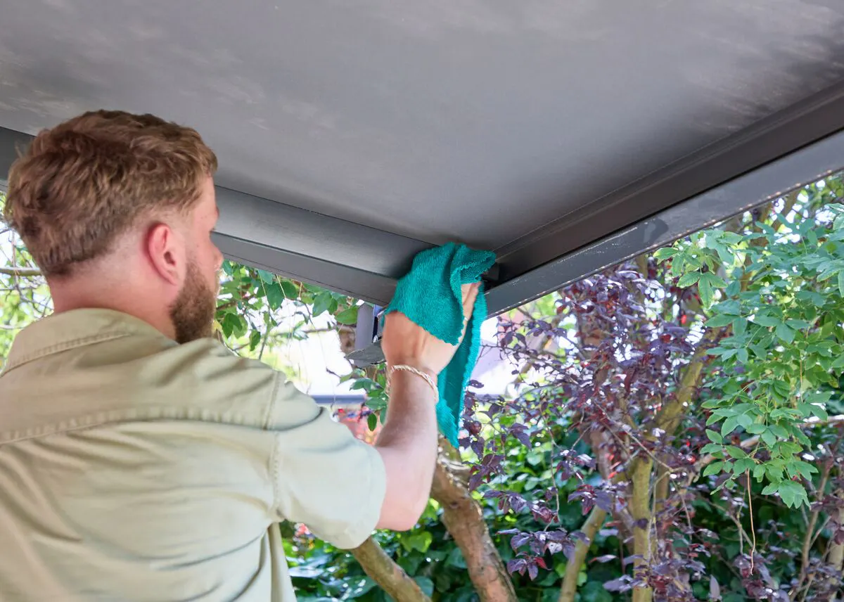 Person wiping the frame of a modern pergola with a cloth, cleaning the underside of the roof beside leafy garden planting.
