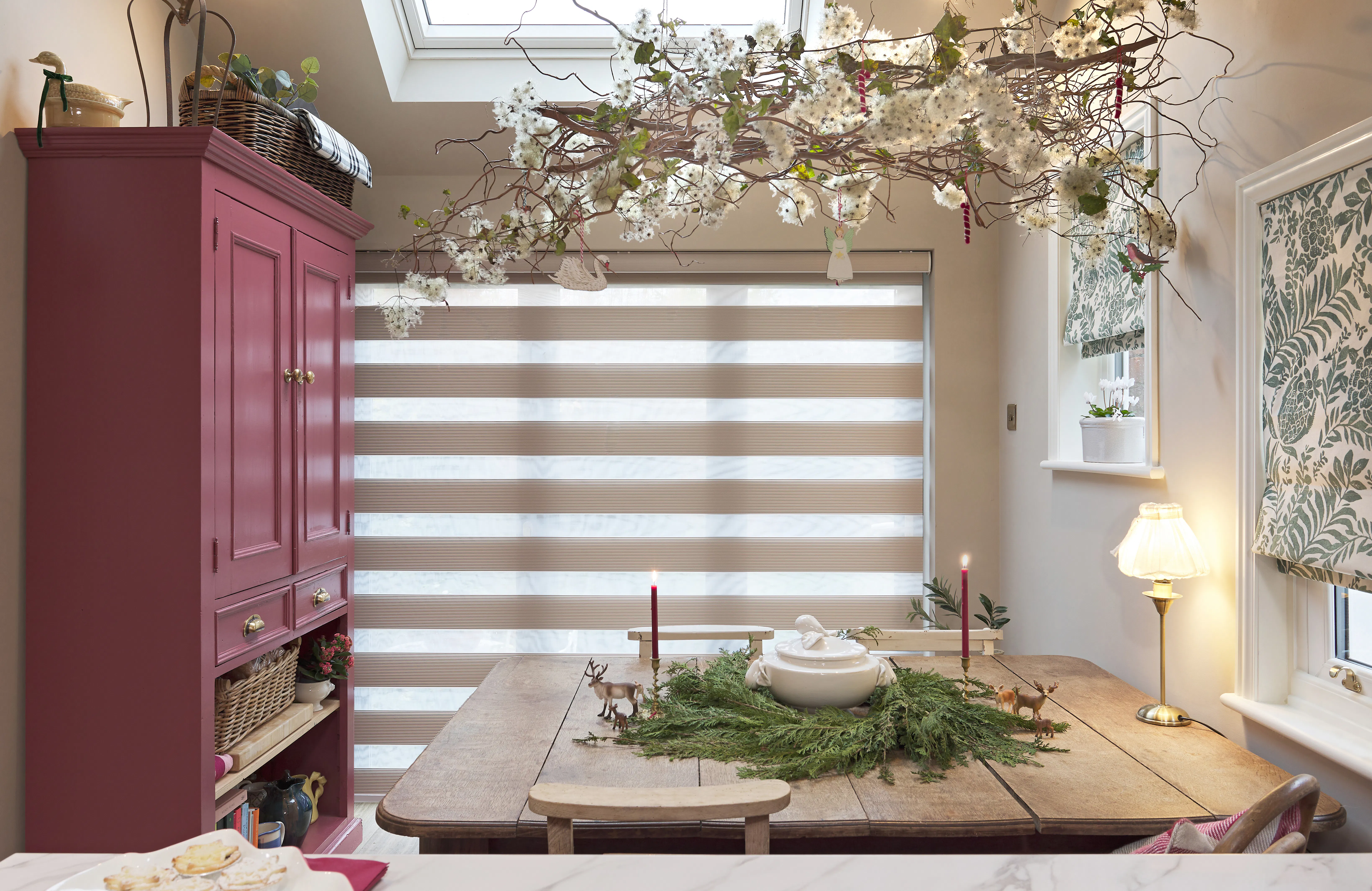 A kitchen dining space with a rose-pink painted cabinet, a striped day-and-night blind at the main window, styled with a wooden table, candles and a blossom branch decoration.