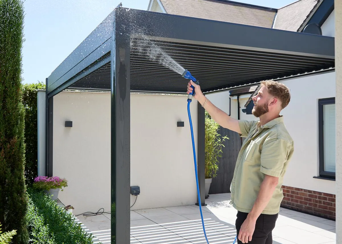 Person rinsing a modern aluminium pergola with a garden hose, washing the frame and louvred roof on a sunny patio.