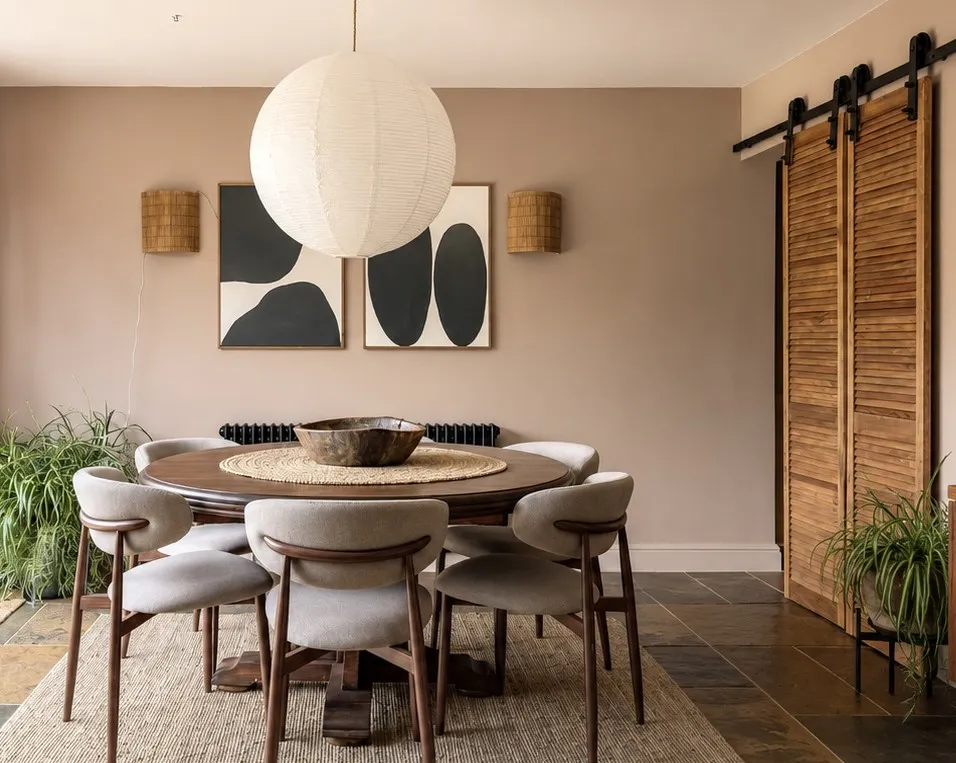 Warm dining room with round wooden table and six upholstered chairs, neutral walls, woven pendant light, abstract art and rustic sliding wooden door.