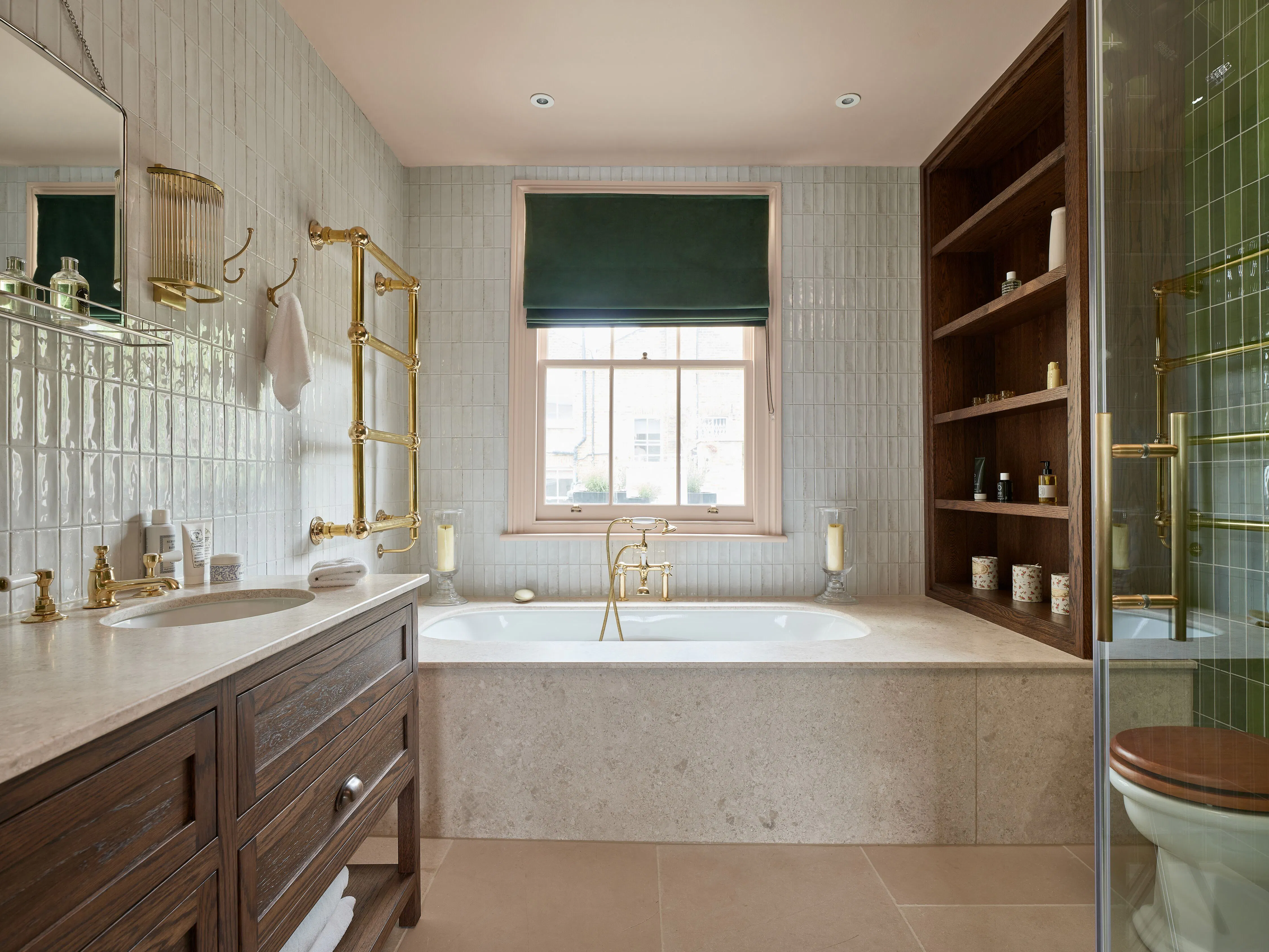 Luxury bathroom with cream tiling, a dark green velvet Roman blind, brass fittings and a built-in wooden shelving unit beside a freestanding bath.
