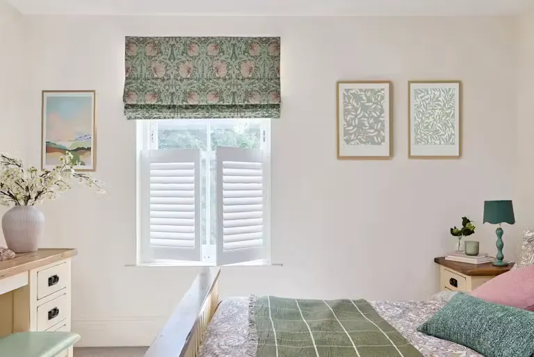 Bedroom window with white shutters and patterned Roman blind, framed by soft décor, wooden furniture and green bedding.