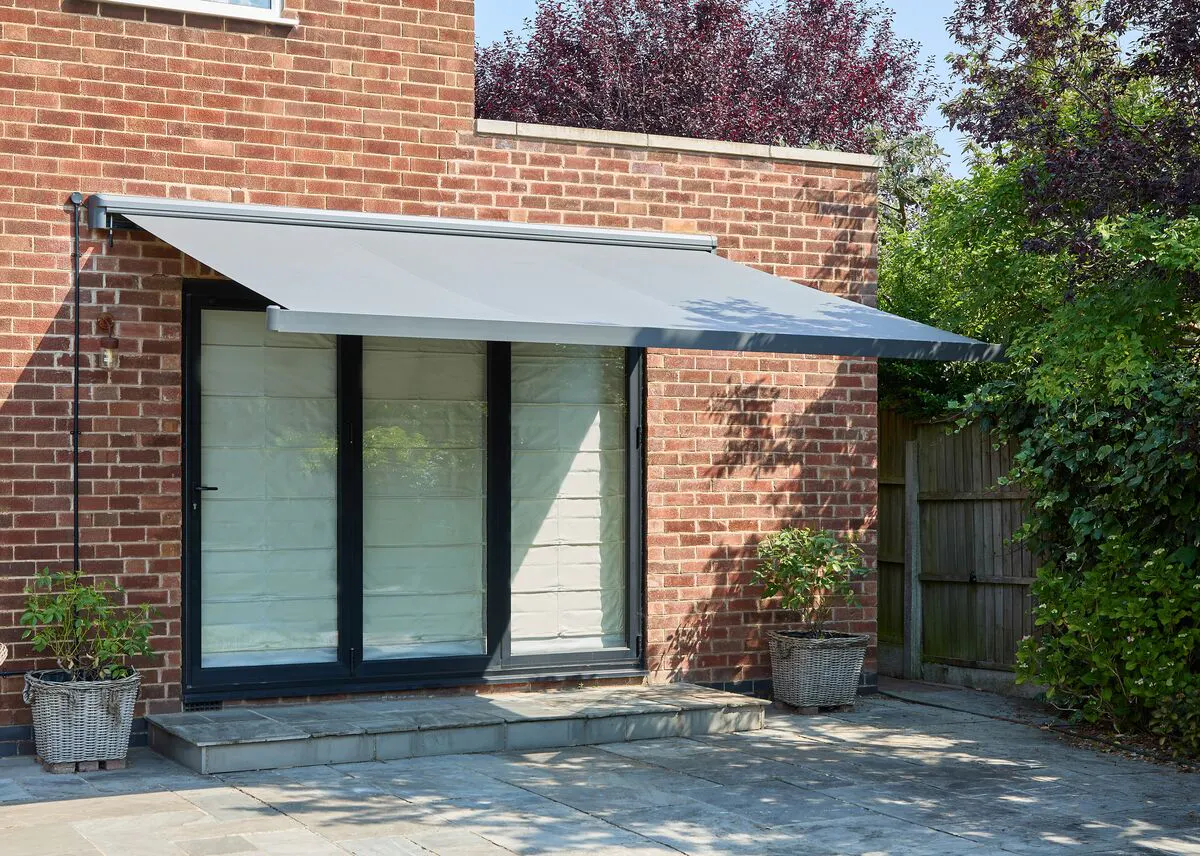 Grey patio awning extended over glazed doors and a paved terrace, providing shade beside a brick house and garden planting.