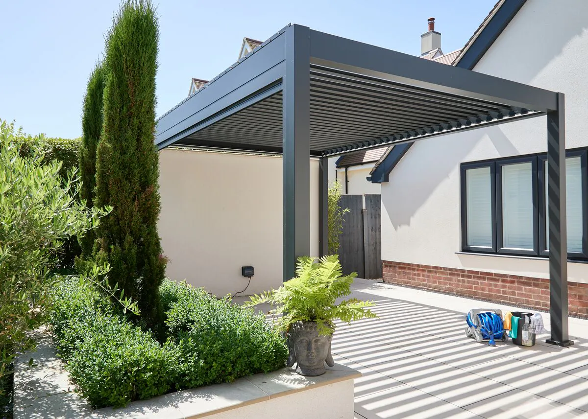 Modern aluminium pergola with a louvred roof shading a contemporary patio, framed by tall greenery beside a white house.