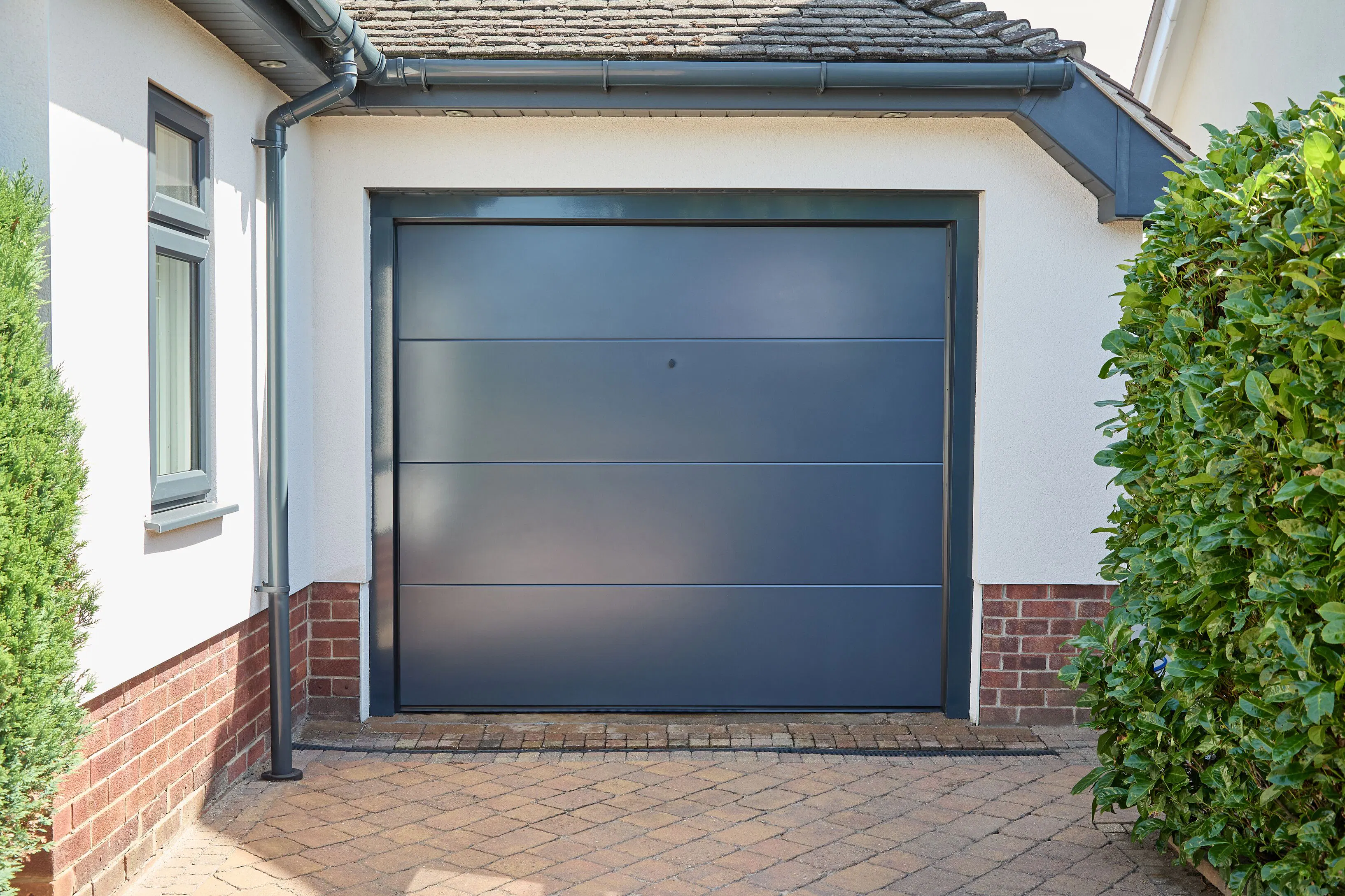 Modern anthracite grey garage door with horizontal panels, framed by white render, brickwork and a front window on a paved driveway.