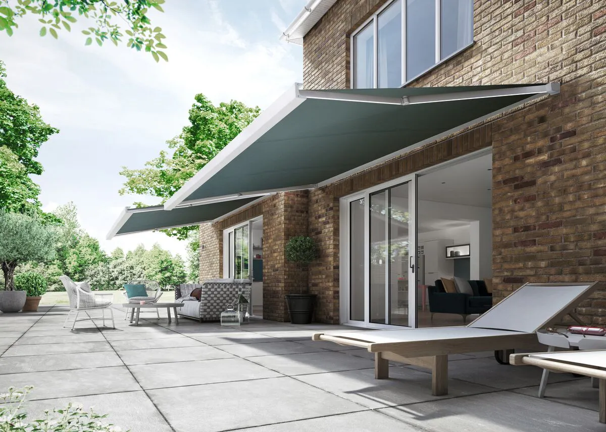 Two grey retractable awnings extended over a spacious patio, shading loungers and seating outside a modern brick house.