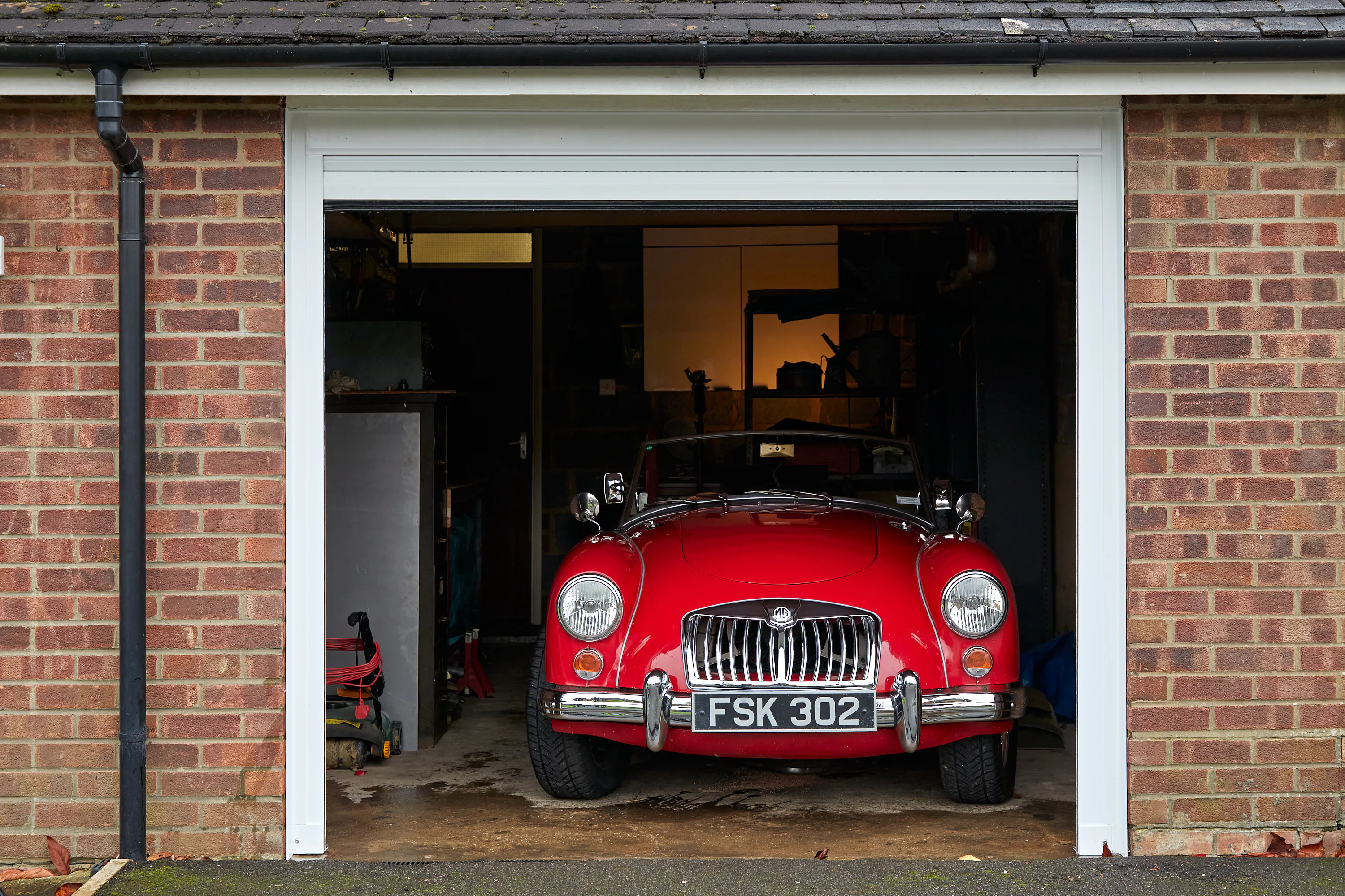 A classic red sports car parked just inside an open garage, framed by a white surround set into a red-brick wall.