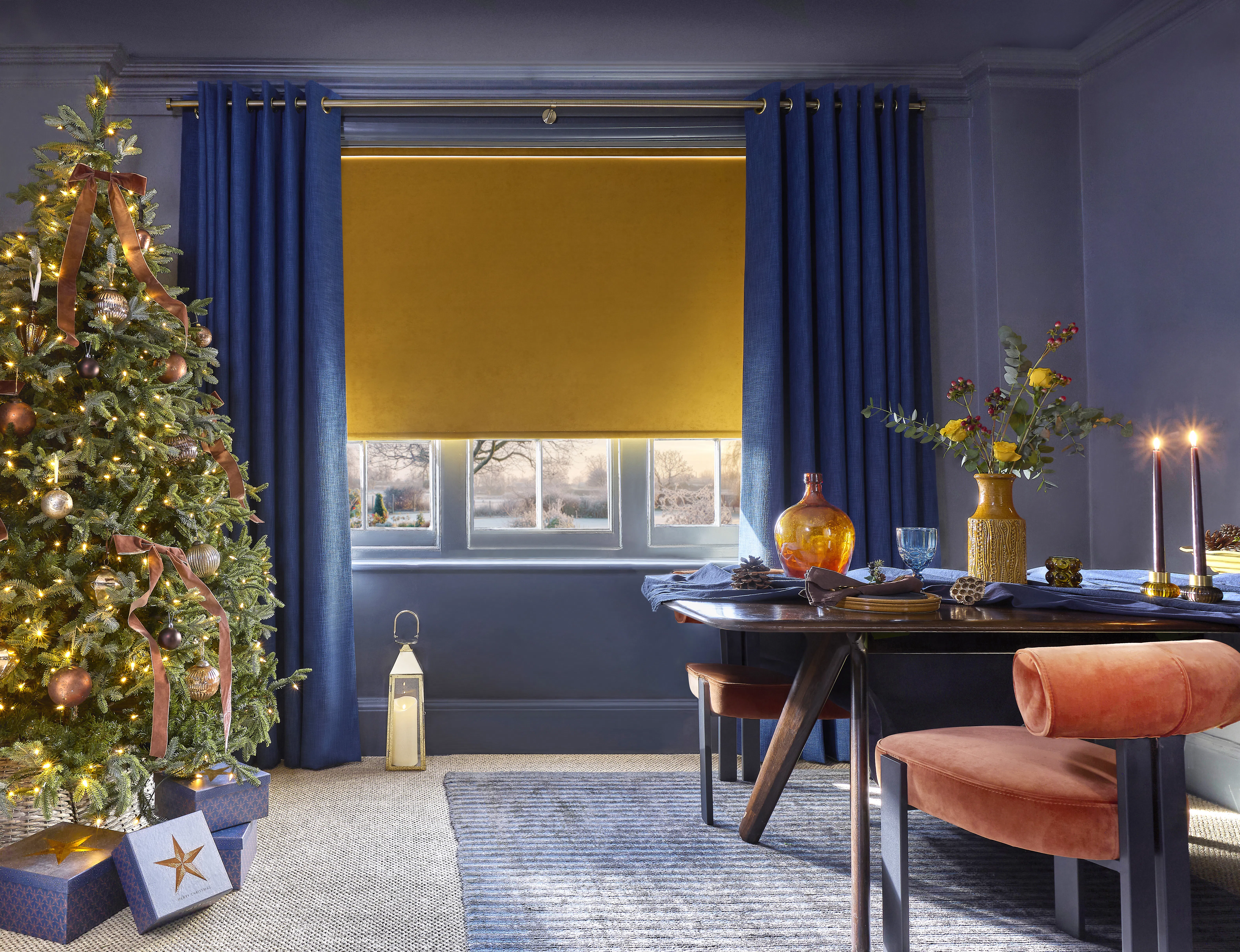 Mustard yellow roller blind with navy blue curtains in a dining room decorated for Christmas, featuring a lit Christmas tree and winter window view.