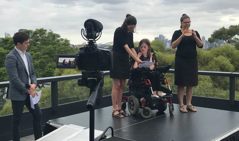 Image of Hireup user, Kate Thomas (centre) on stage joined by Jordan O'Reilly to her right and an AUSLAN interpreter to the left.