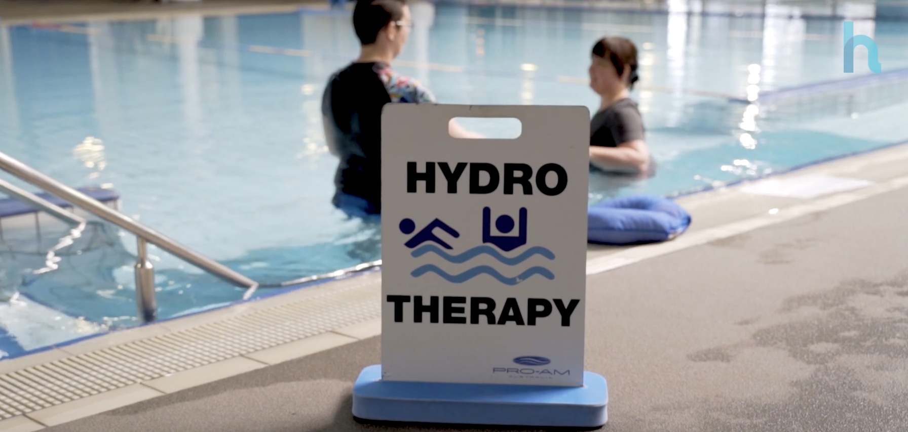 Hydrotherapy sign sits on pool deck as two people use the pool in the background.