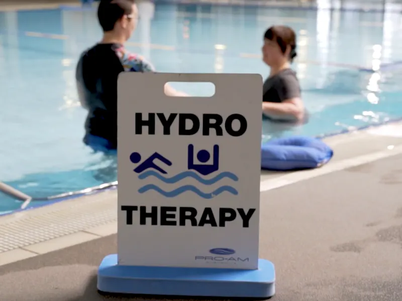 Hydrotherapy sign sits on pool deck as two people use the pool in the background.