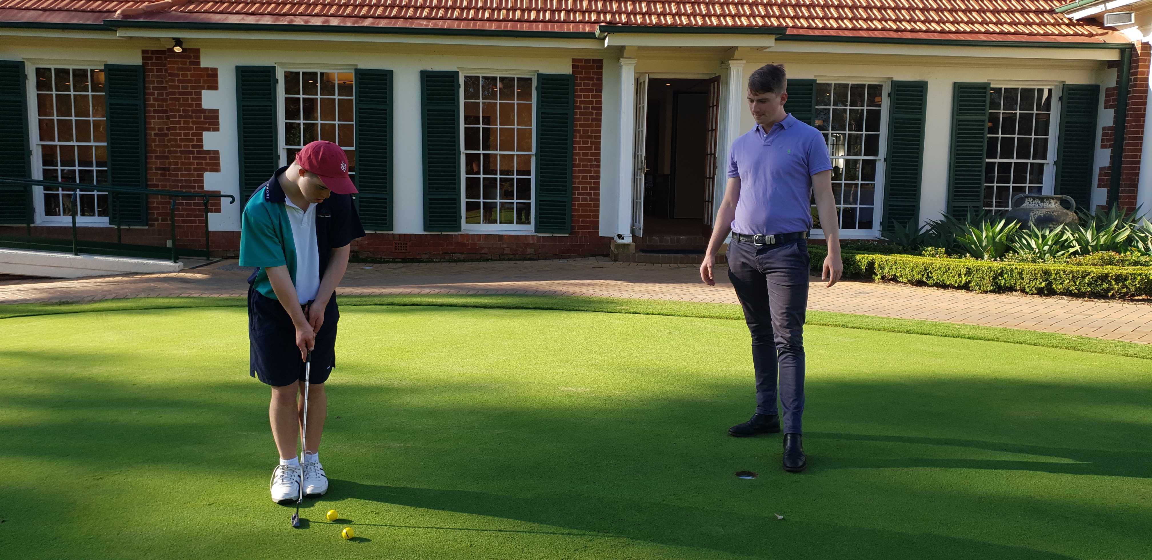 Jamie and Pat practice their golfing skills on a green in front of a clubhouse.