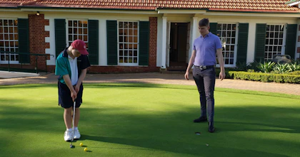 Jamie and Pat practice their golfing skills on a green in front of a clubhouse.