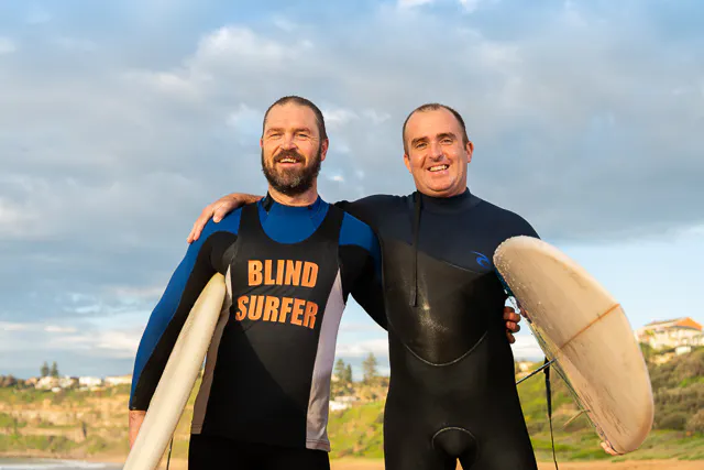 Hireup users Kirk (left) and Dan (right) stand arm in arm as they pose for the camera after their morning surf.