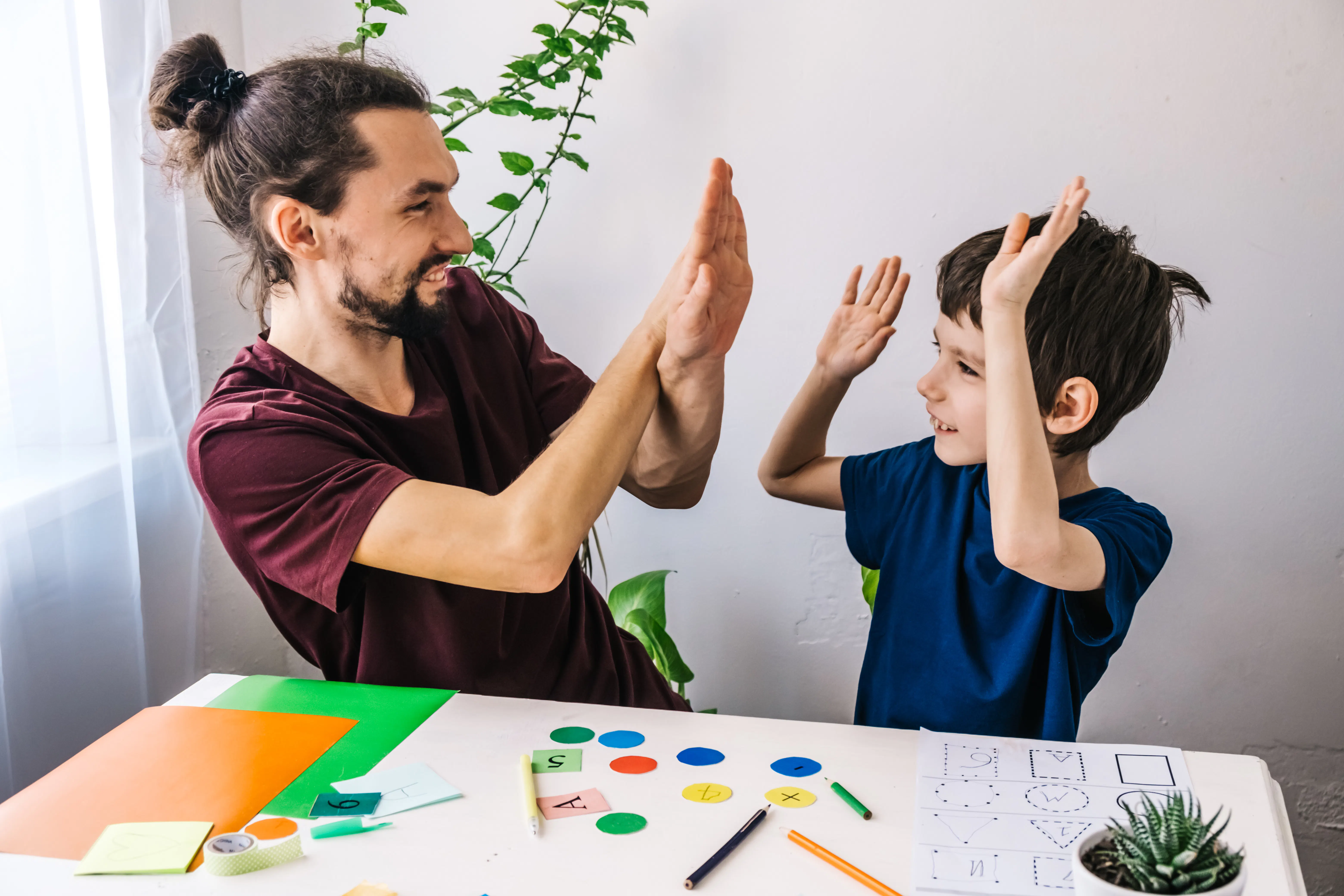 A young man and young boy are sitting beside each other at a desk giving each other a double hi-five. The man has long dark brown hair tied up in a bun. He has facial hair and is wearing a maroon t-shirt shirt. The boy has short brown hair and is wearing a dark blue t-shirt. There are coloured pencils and paper on the table in front of them.