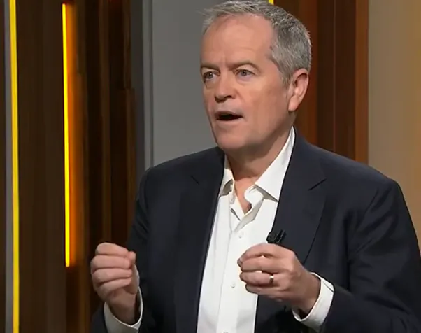 Bill Shorten is talking. He wears and white shirt and dark suit jacket. His hands are raised.  