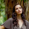 Autistic writer and advocate Amy Cramb, a woman with olive skin, long brown hair and dark brown eyes, stands in front of a tree in a green, leafy setting. Amy wears a brown silky top with short, billowy leaves. She appears calm and content.