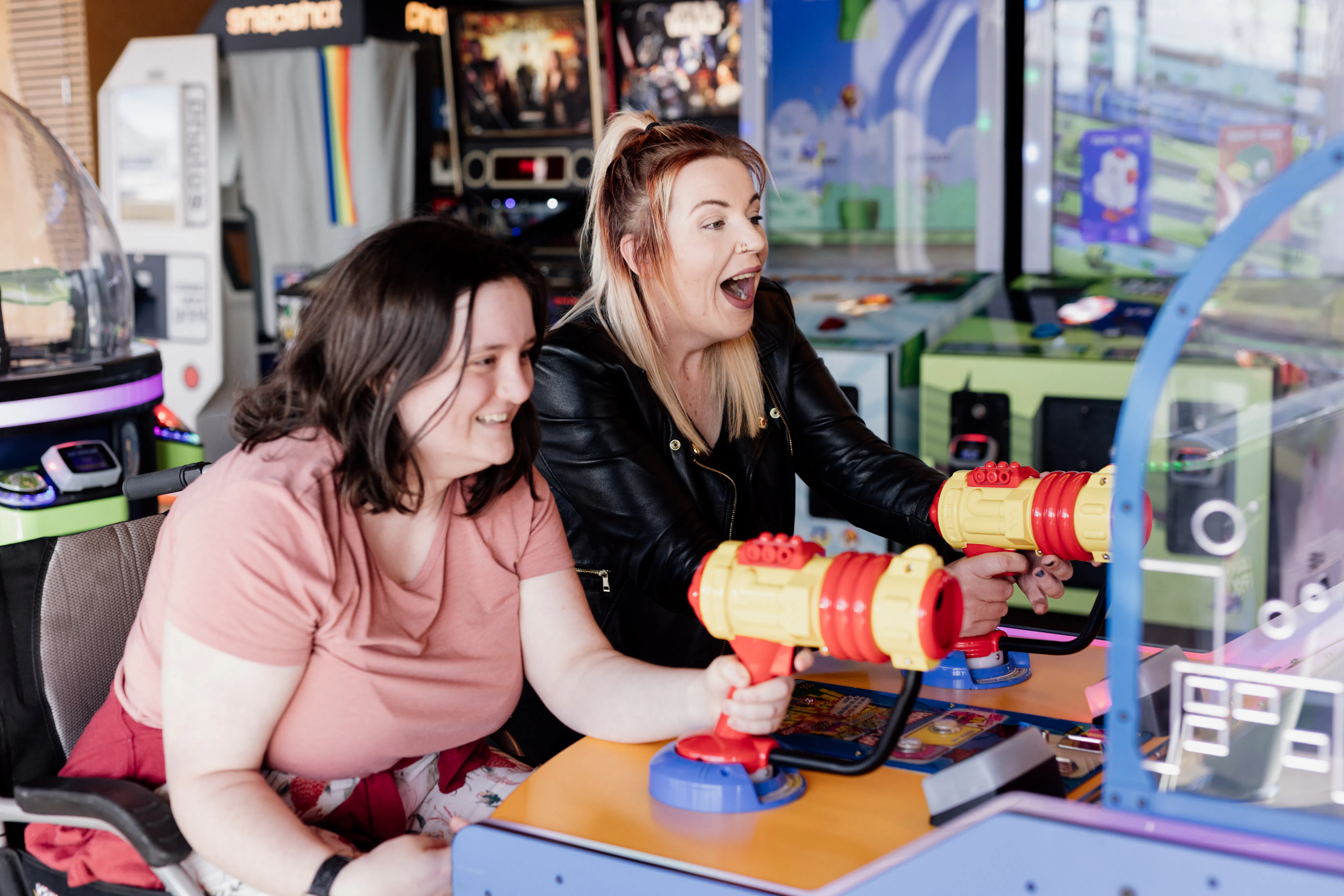 Hayley and Rebekah are playing a shooting game at an arcade. They are both smilng. Hayley has dyed blonde hair and is wearing a black leather jacket and a black t-shirt. Rebekah has shoulder length brown hair and is wearing a pink t-shirt.