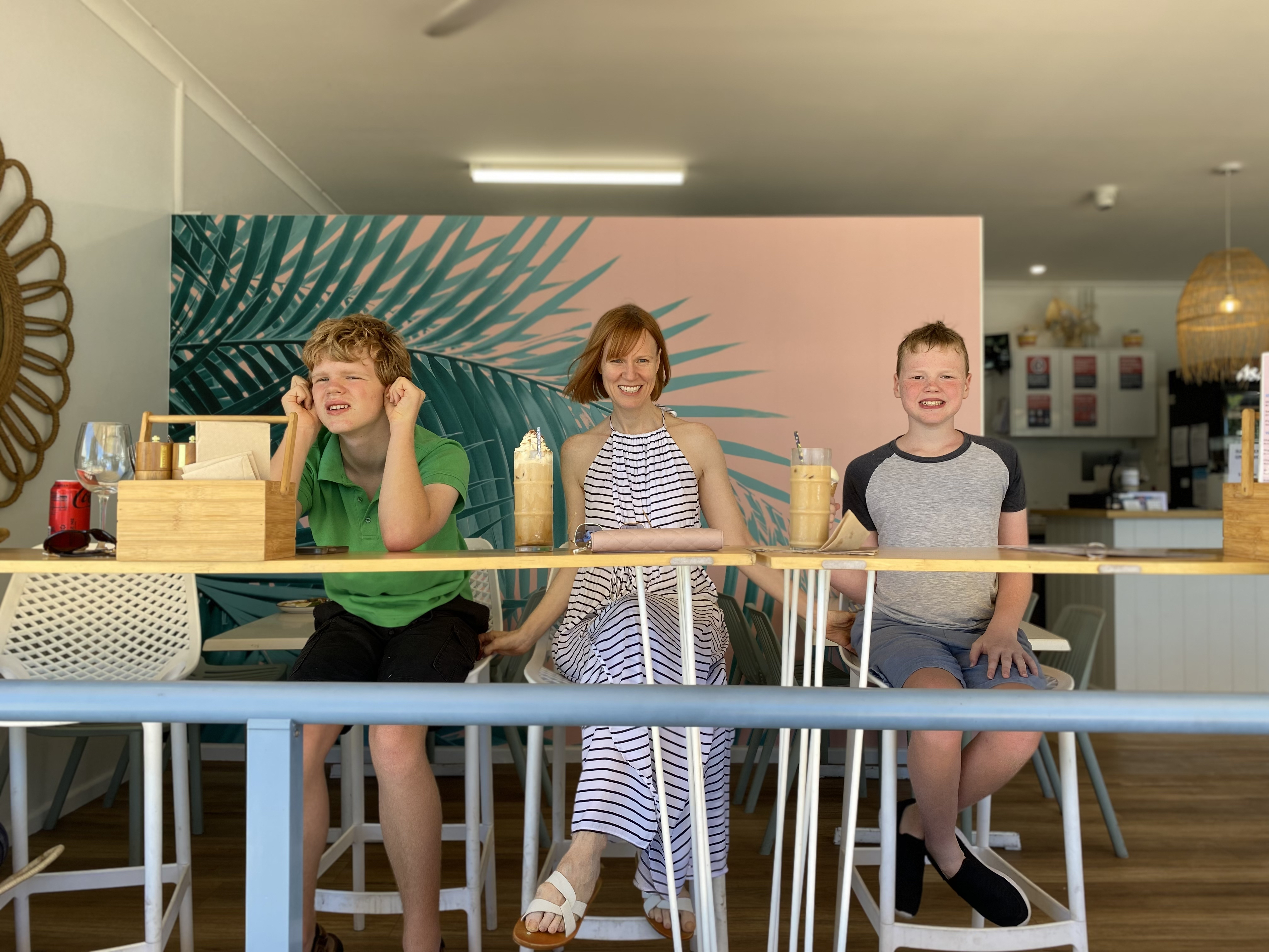 Briana Blackett, a woman with red hair wearing a white and black-striped halter dress, sits smiling between her two sons. The trio are at a cafe drinking ice chocolates. 