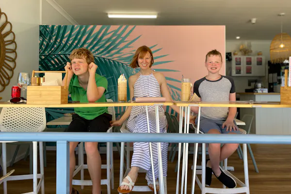 Briana Blackett, a woman with red hair wearing a white and black-striped halter dress, sits smiling between her two sons. The trio are at a cafe drinking ice chocolates.