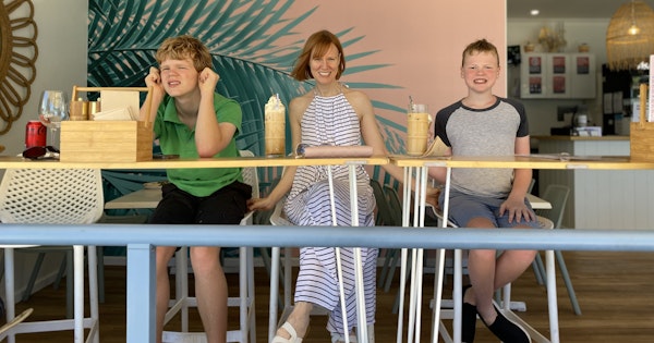 Briana Blackett, a woman with red hair wearing a white and black-striped halter dress, sits smiling between her two sons. The trio are at a cafe drinking ice chocolates.