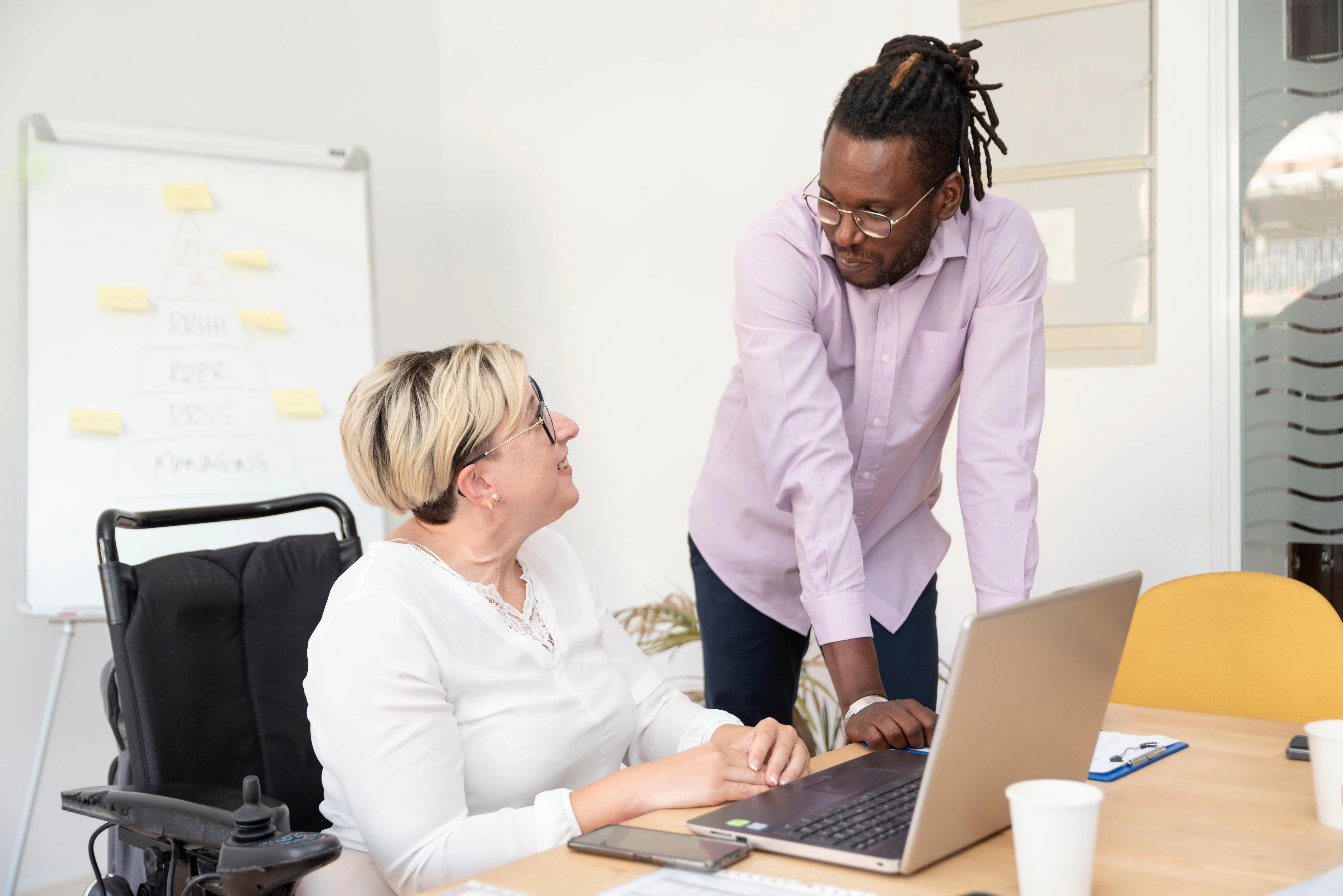 A woman with short dyed blonde hair is sitting at a desk with a laptop. She is wearing a white long sleeve top and black rimmed glasses. She is smiling at a man next to her. The man has black dreadlocks and is wearing a light purple collared shirt and black pants. The woman is a wheelchair user. 