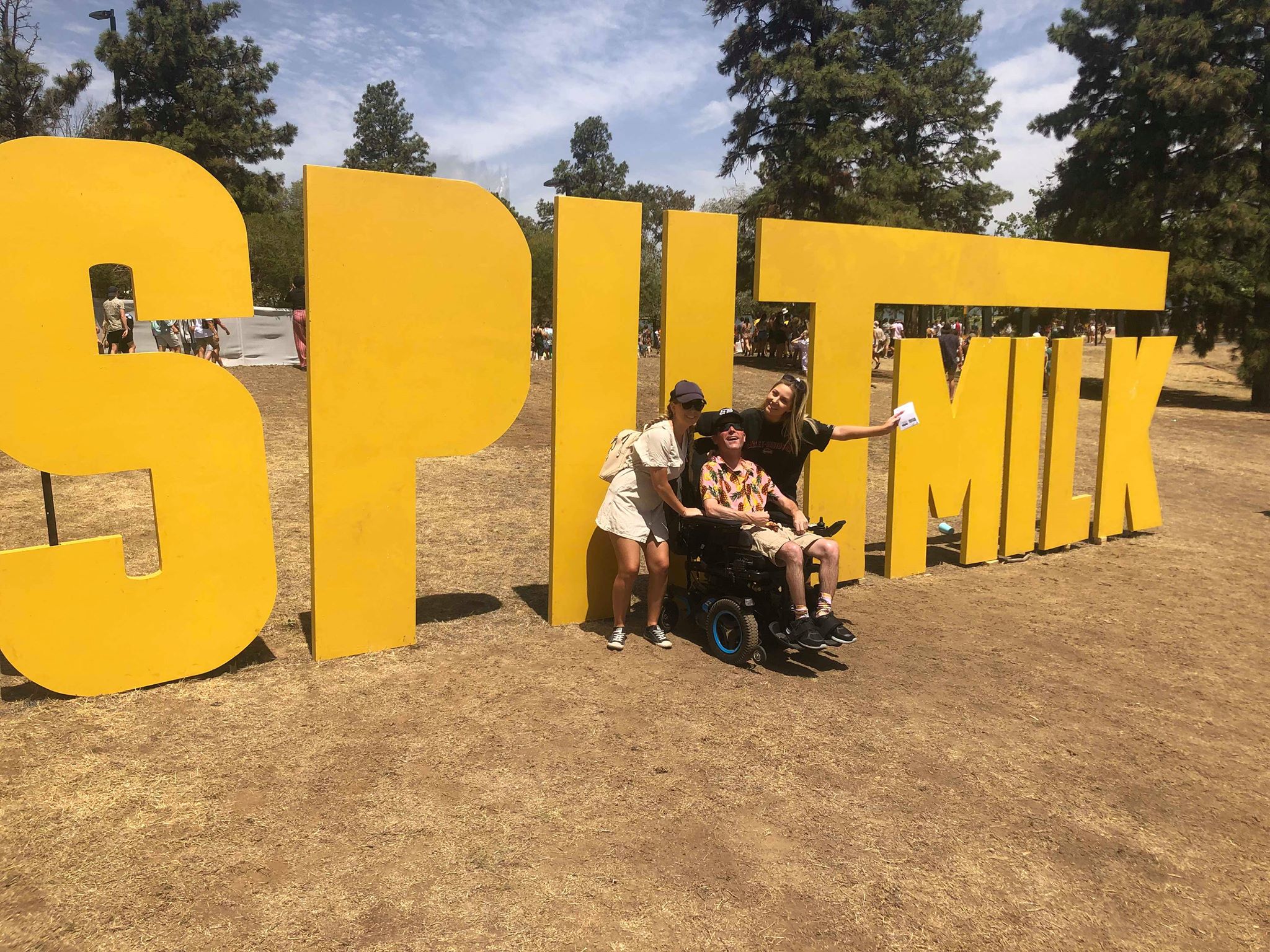 Mick is seated in his wheelchair next to two of his support workers. They are posing in front of the large Spilt Milk festival sign.
