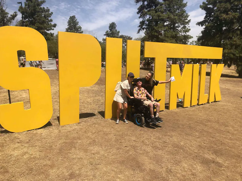 Mick is seated in his wheelchair next to two of his support workers. They are posing in front of the large Spilt Milk festival sign.