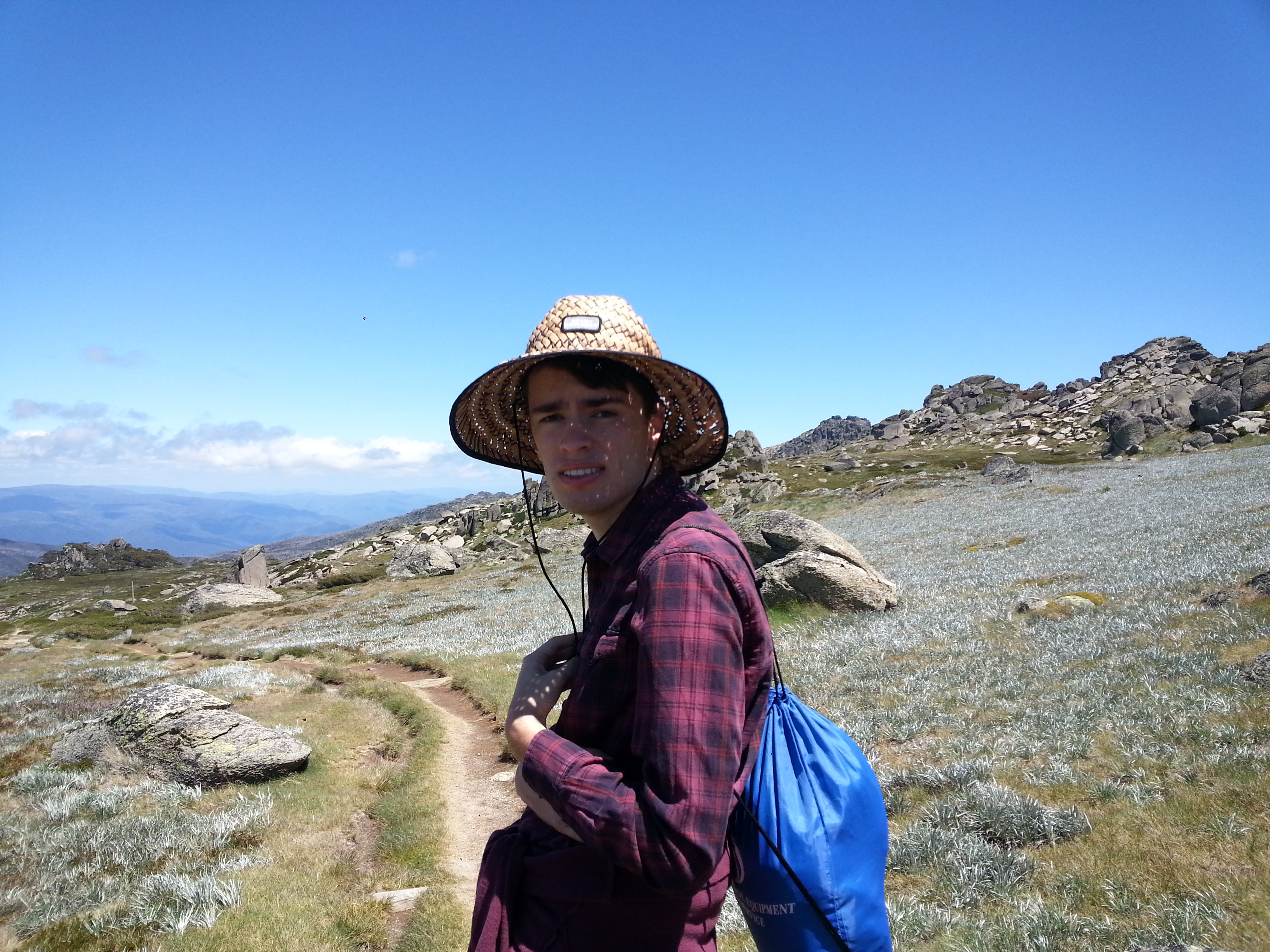 Andrew is on a walking track. He is wearing a large straw hat and a red flannelette shirt. 