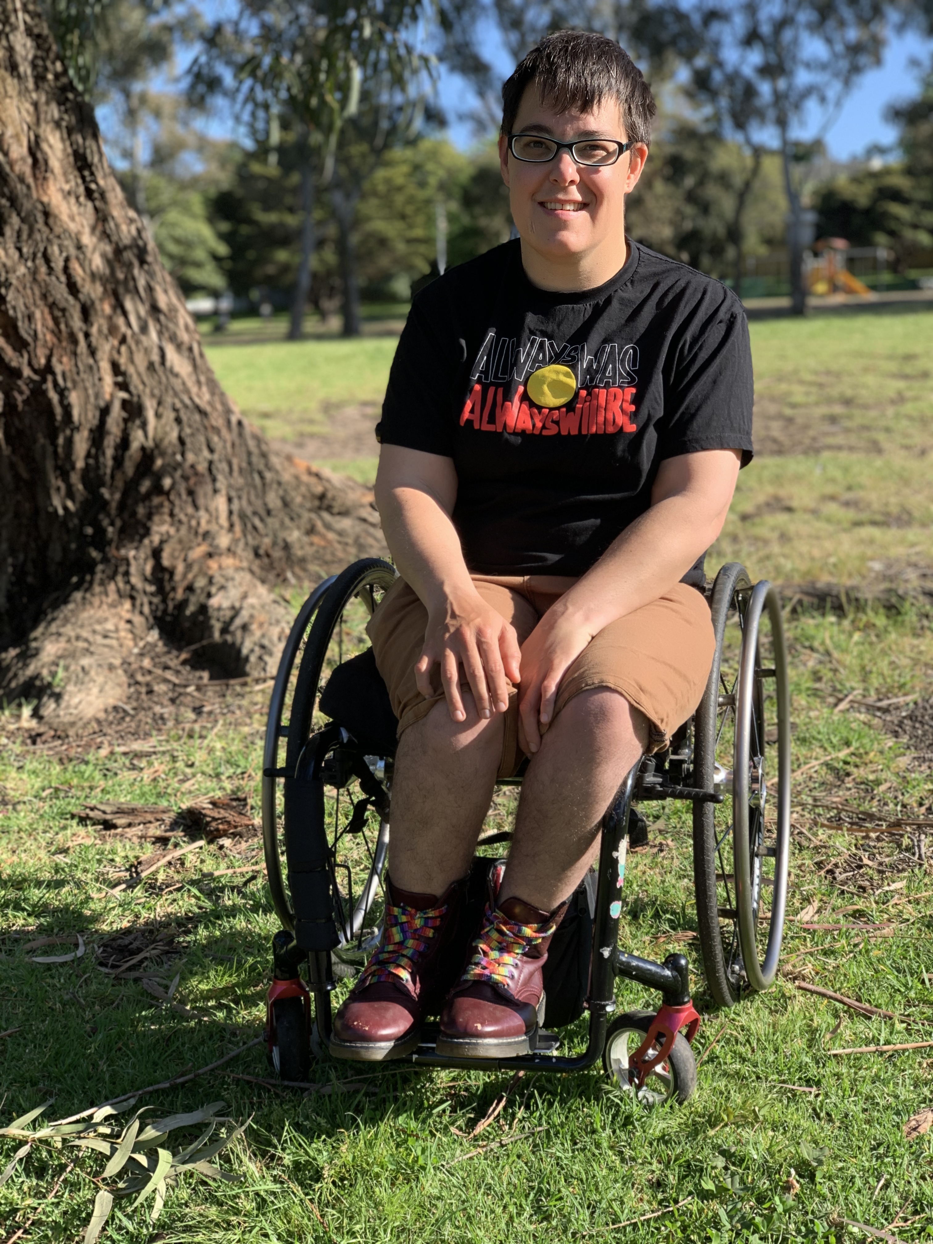 Jax is wearing a dark shirt, light shorts, and shoes with rainbow laces. They're in a park and are smiling. Jax is a wheelchair user. 