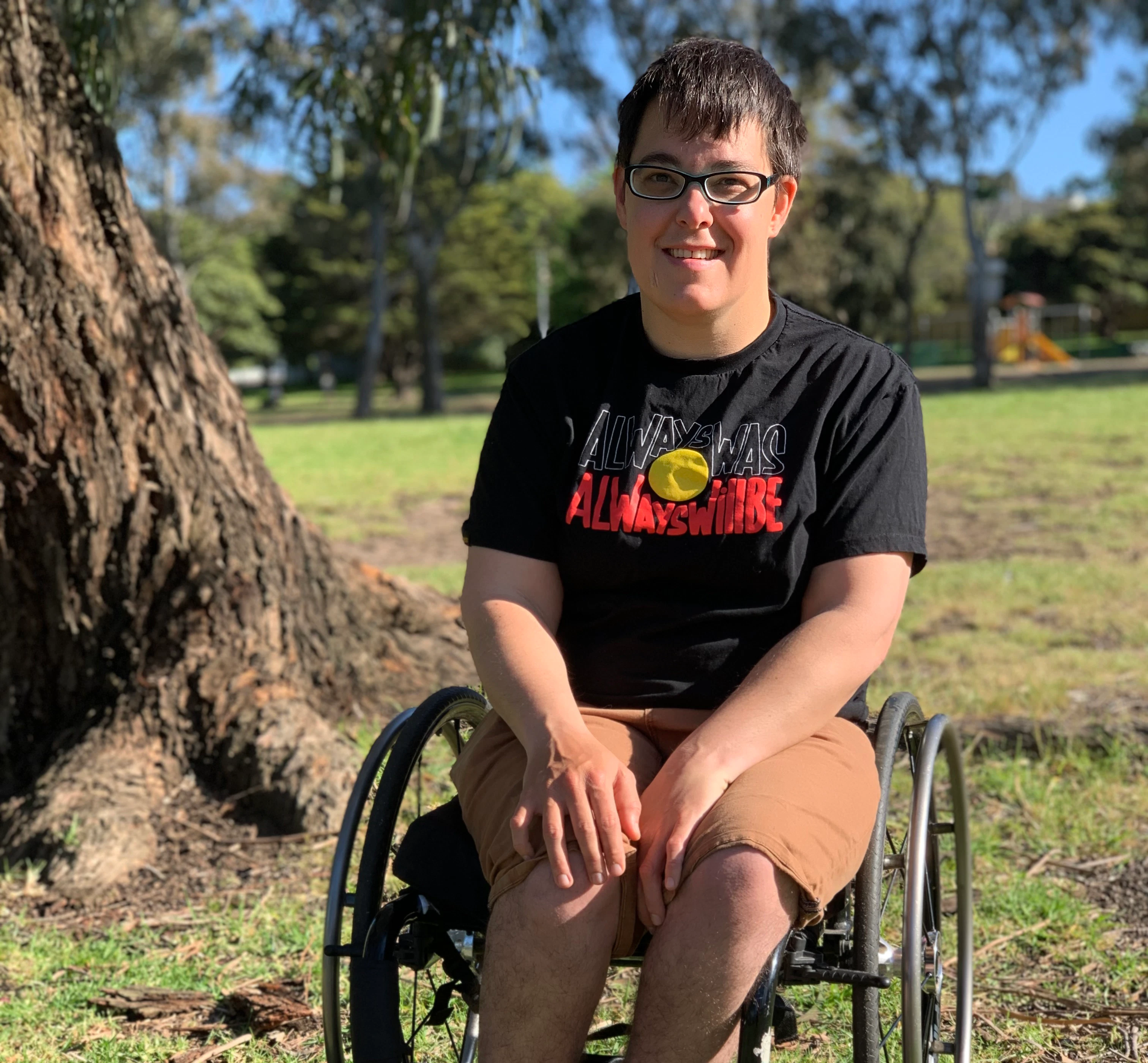 Jax is wearing a dark shirt, light shorts, and shoes with rainbow laces. They're in a park and are smiling. Jax is a wheelchair user.