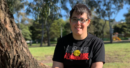 Jax is wearing a dark shirt, light shorts, and shoes with rainbow laces. They're in a park and are smiling. Jax is a wheelchair user.
