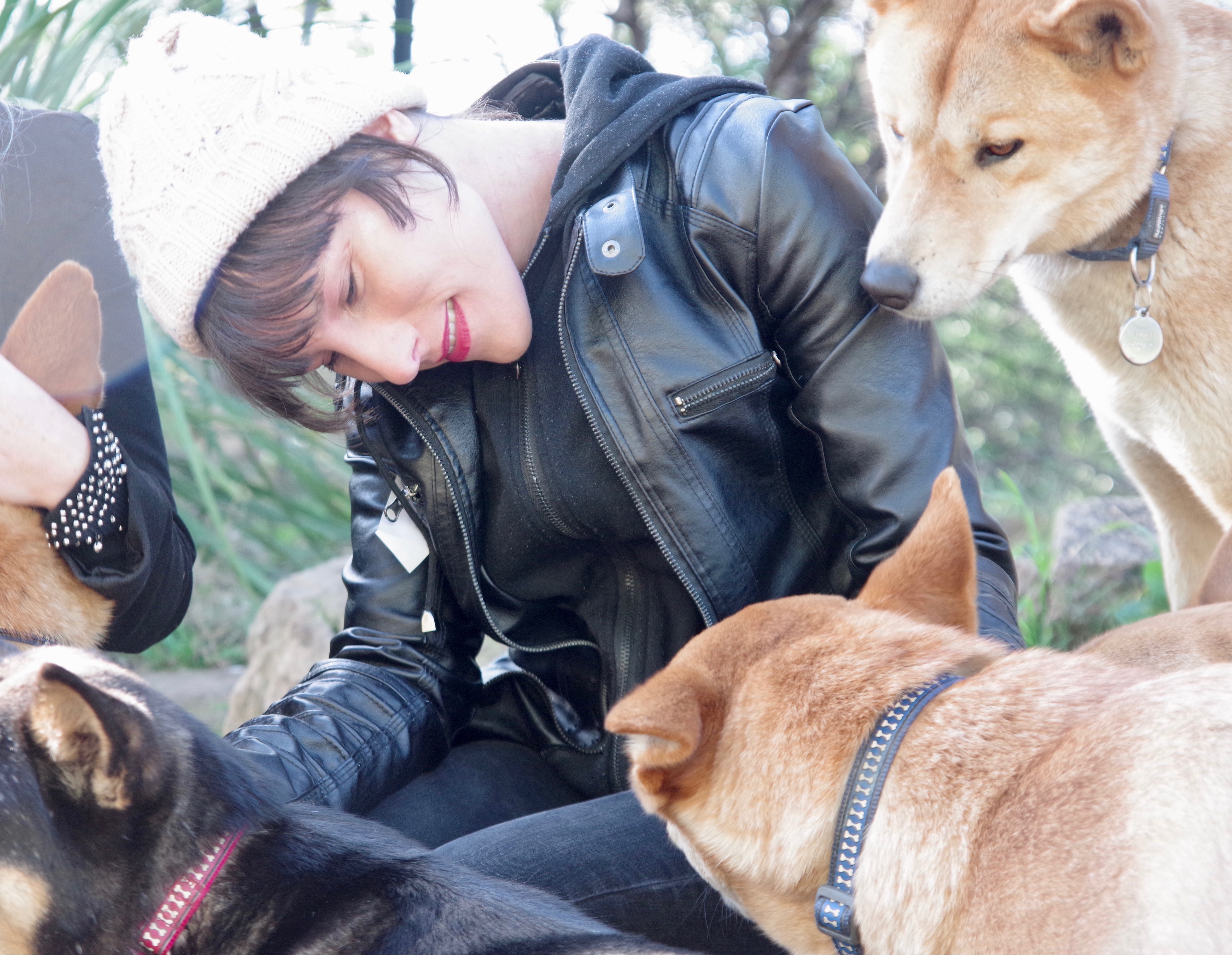 Elena Wewer, a woman with dark brown hair, white skin and bright pink lipstick, looks loving at a group of dogs that surround her. Elena wears a black leather jacket and white knitted beanie. Two of the dogs are caramel coloured, while another is tan and black.  