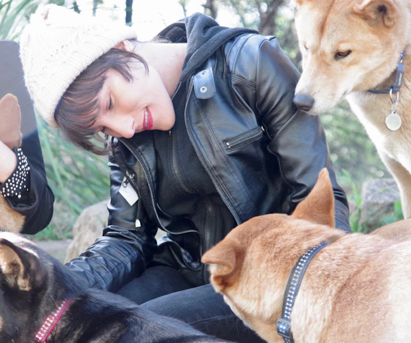 Elena Wewer, a woman with dark brown hair, white skin and bright pink lipstick, looks loving at a group of dogs that surround her. Elena wears a black leather jacket and white knitted beanie. Two of the dogs are caramel coloured, while another is tan and black.