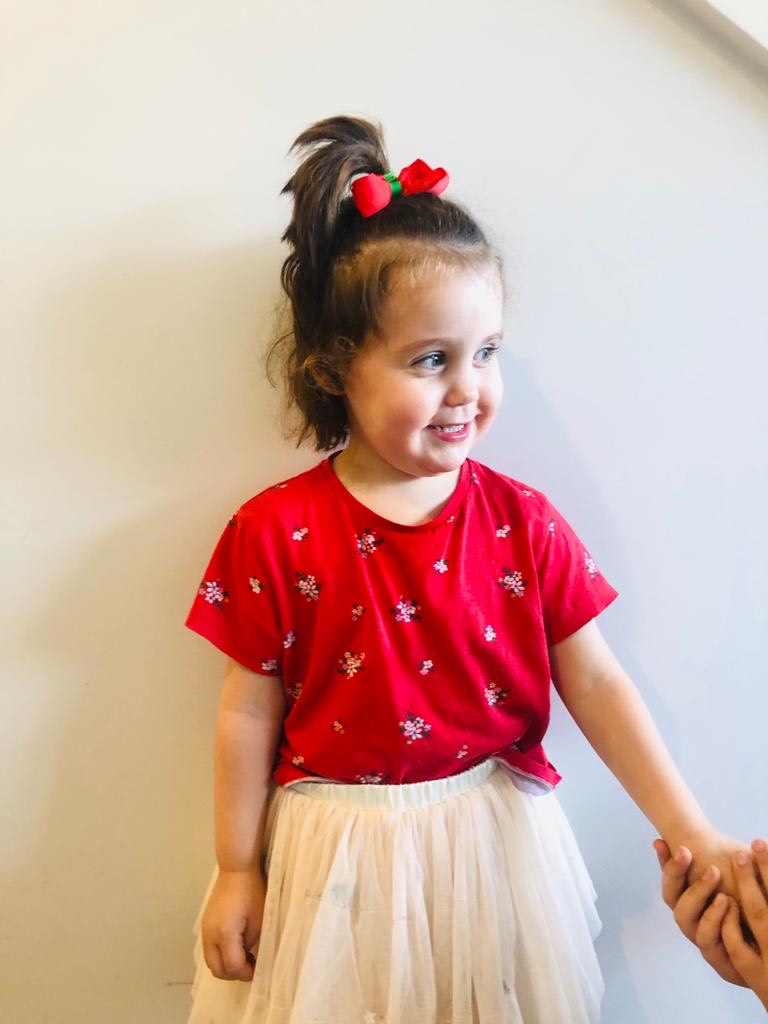 Coco is wearing a red shirt, a ballerina tutu, and a red ribbon in her hair to match. She is looking to the side, smiling, and is holding someone's hand. 