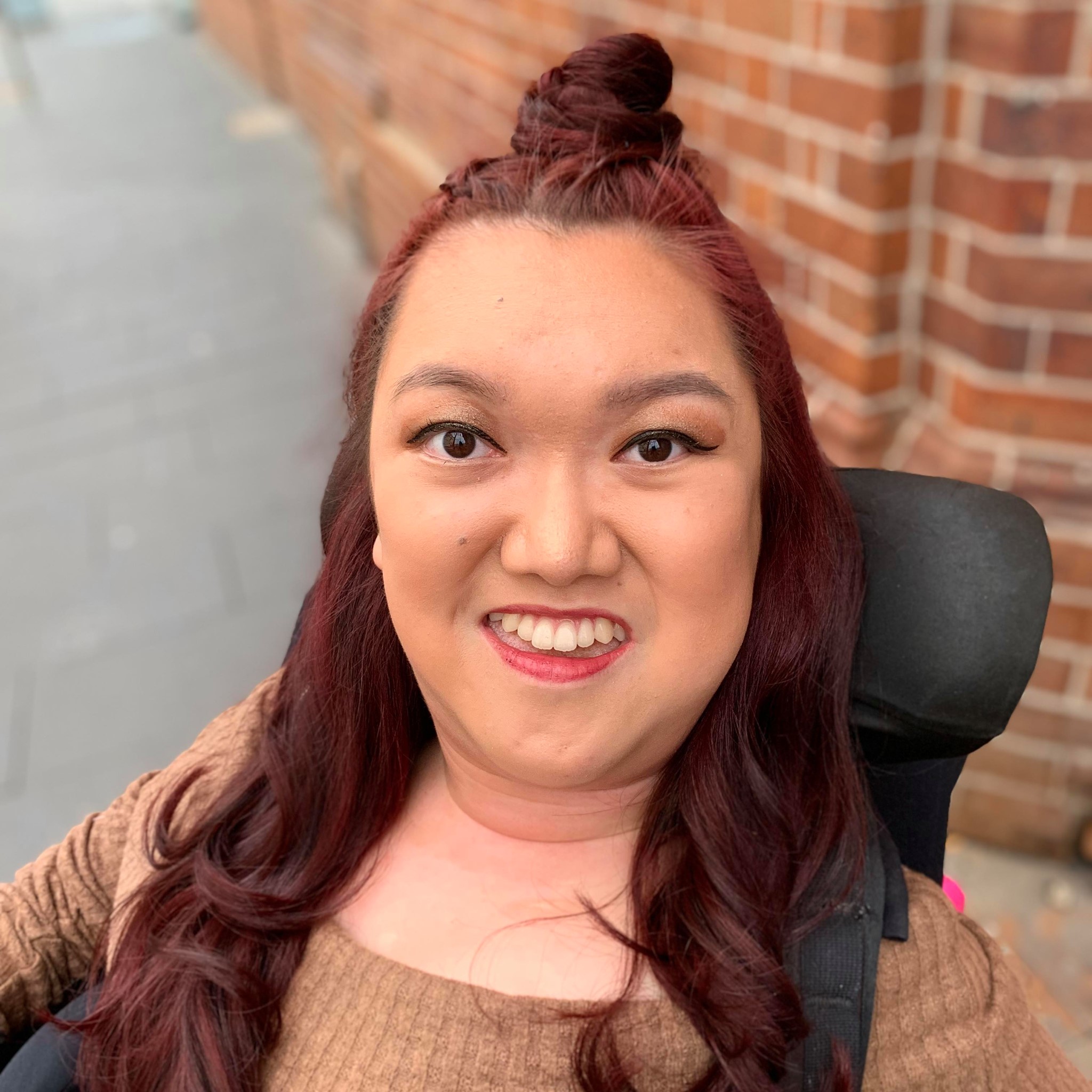 Melanie Tran is smiling. She's on a pathway and a brick building is behind her. Melanie's hair is half up and half out. Melanie is a wheelchair user. 