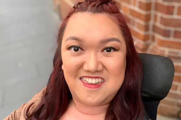 Melanie Tran is smiling. She's on a pathway and a brick building is behind her. Melanie's hair is half up and half out. Melanie is a wheelchair user.