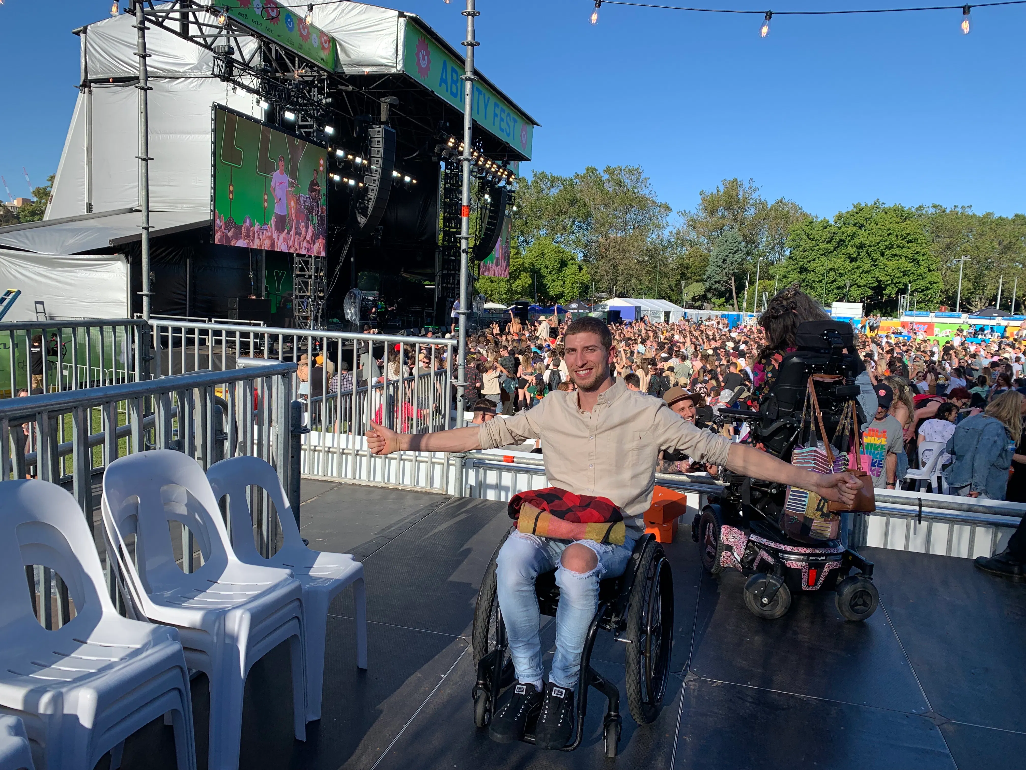 Jacob Darkin has his arms stretched wide and is smiling. He's on the wheelchair platform at a music festival. A crowd of people are in front of the main stage.