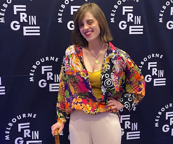 Liel Bridgford has shoulder length brown hair and wears a colourful shirt while posing in front of a Melbourne Fringe Festival banner. She holds a walking cane and smiles at the camera.